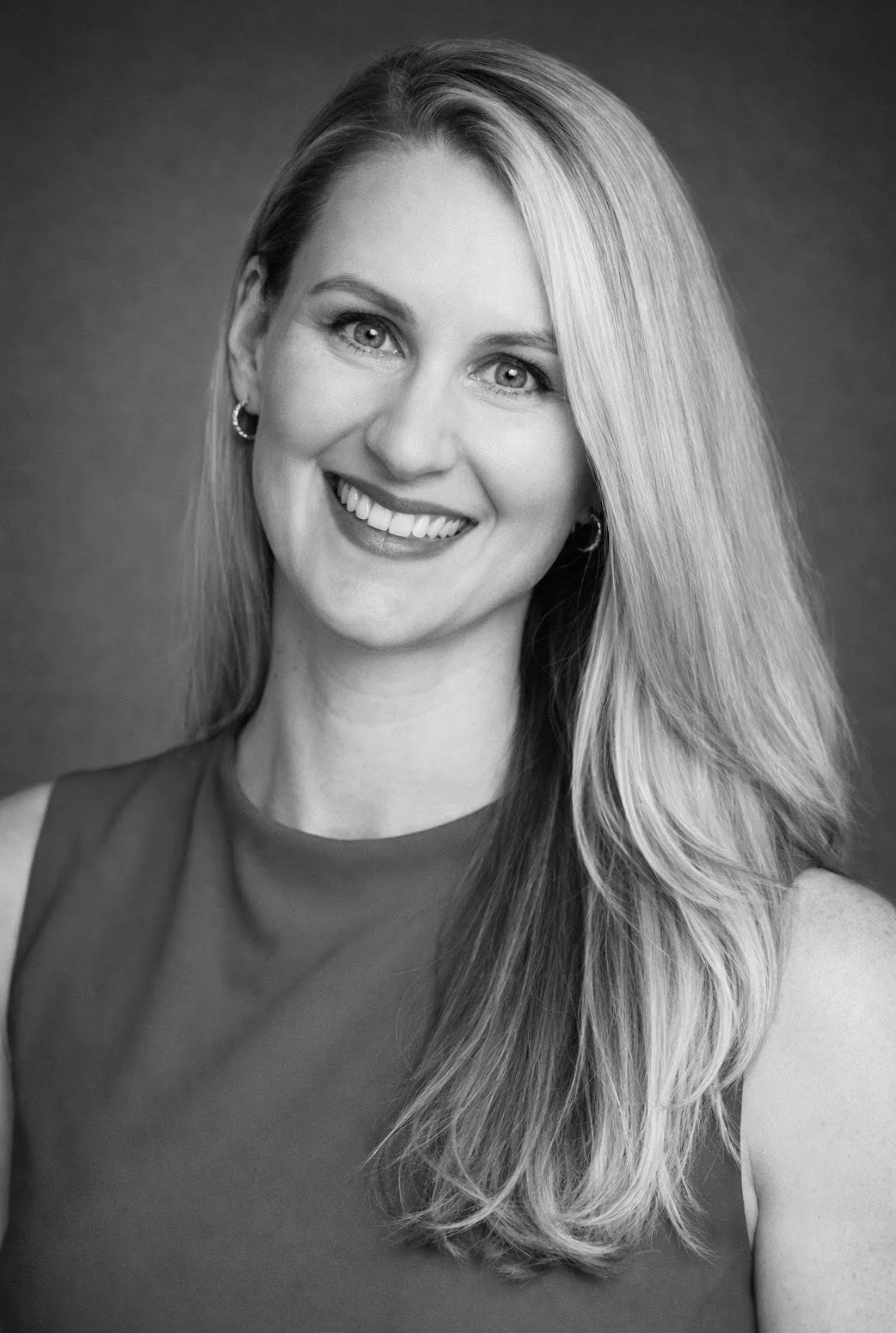 Black and white portrait of a woman with long blonde hair, smiling, wearing earrings and a sleeveless top.