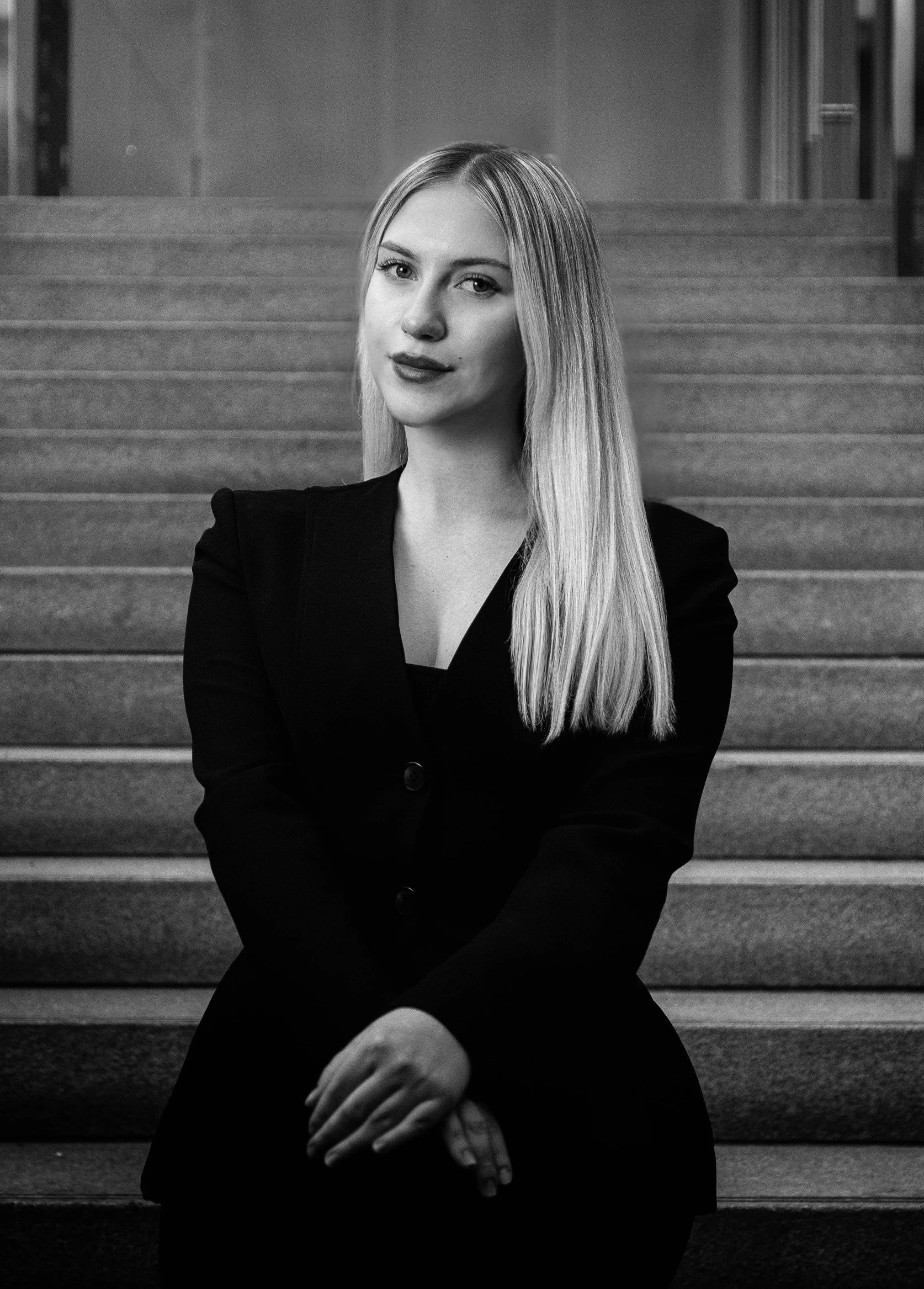 A black and white photo of a woman with long, straight hair sitting on stairs, looking directly at the camera with a slight smile, wearing a dark blazer.