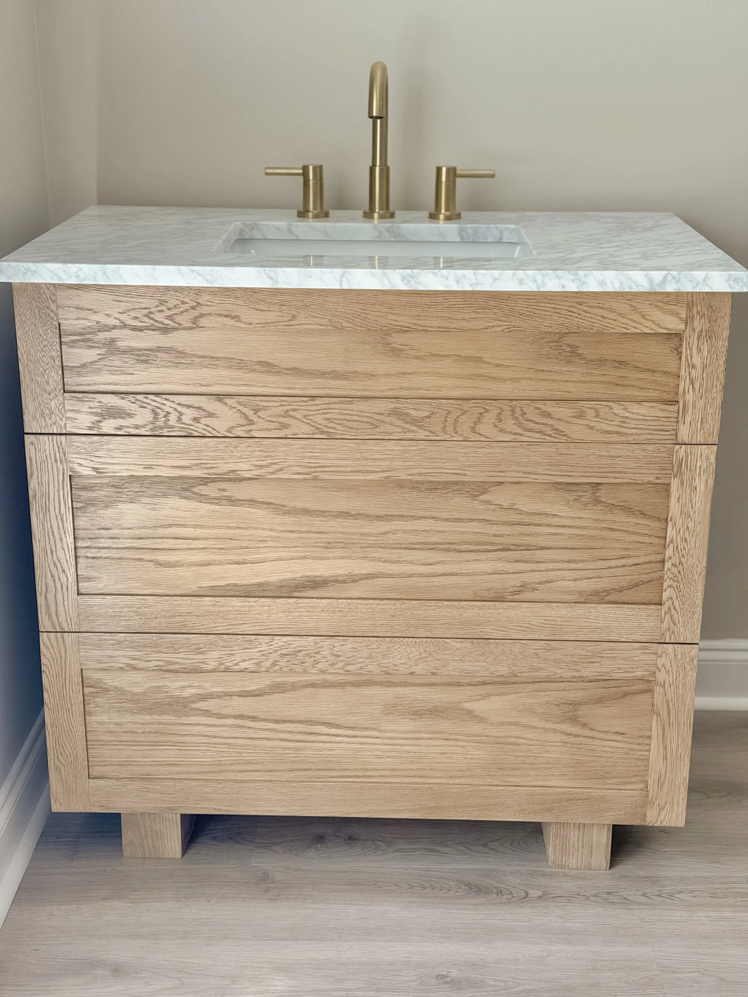 A wooden vanity with a light-colored marble top and gold fixtures, including a faucet and handles, set against a plain wall.