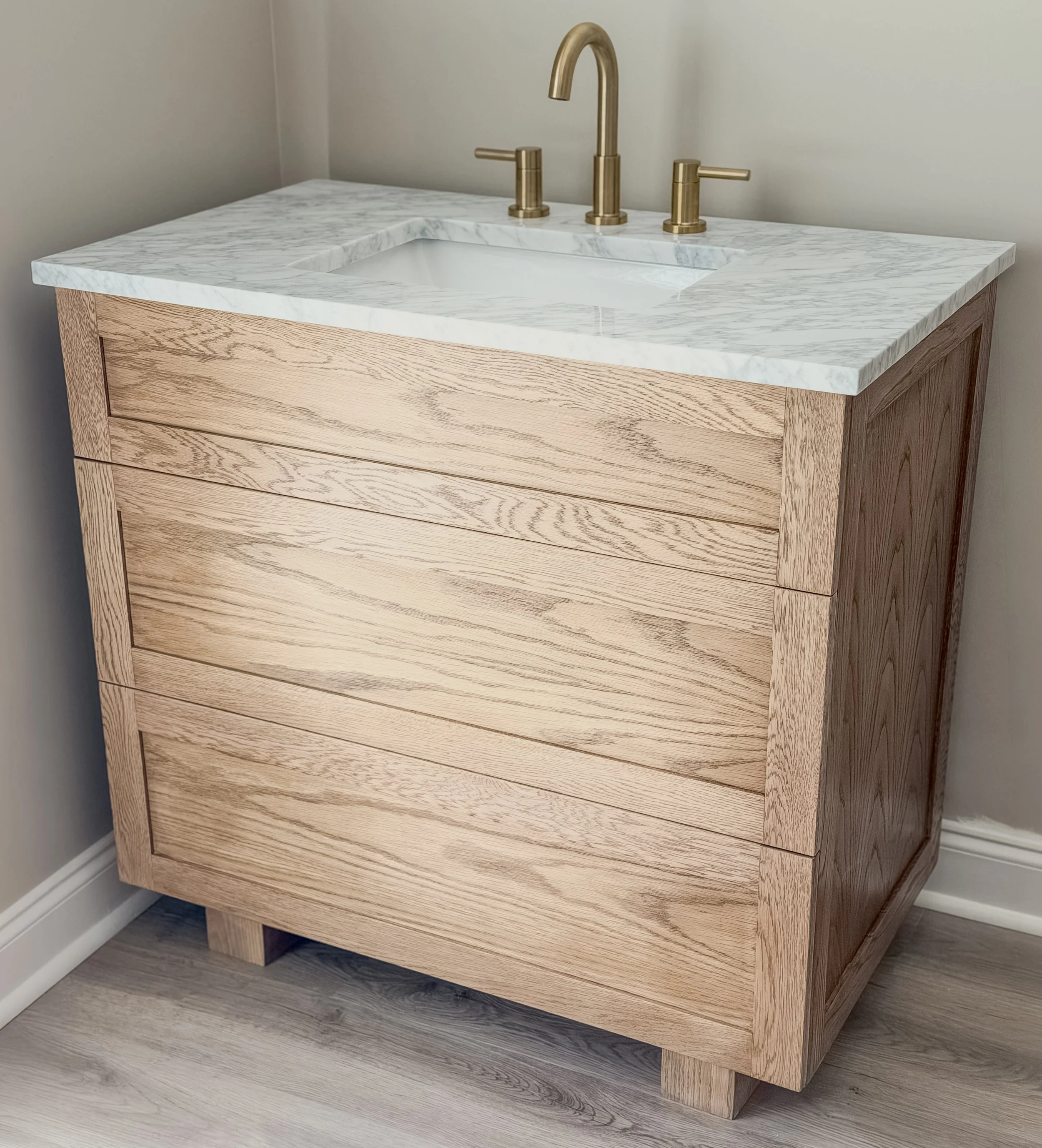 A wooden vanity with a white marble countertop and an integrated sink, featuring a brass faucet and matching brass handles, positioned against a light-colored wall with wood flooring.