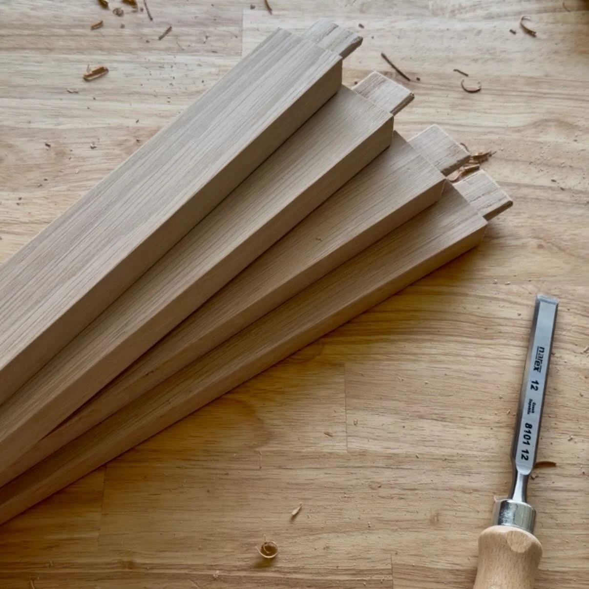 A row of five wooden planks on a wooden surface with sawdust and wood shavings nearby, and a hand tool with a metal blade and wooden handle nearby.