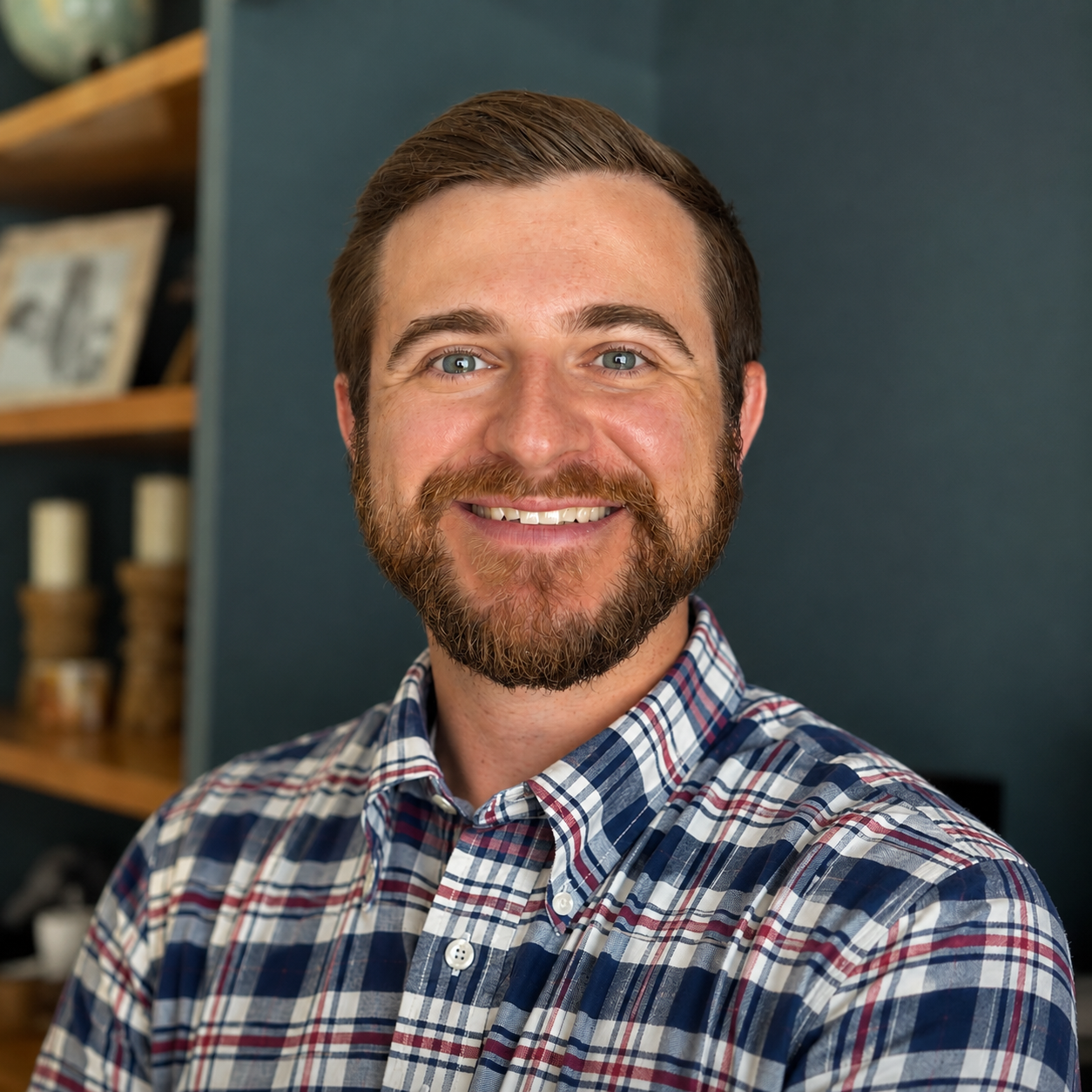 Close-up of smiling man with beard and blue eyes, wearing a plaid shirt, in a room with dark blue walls and bookshelf in background.