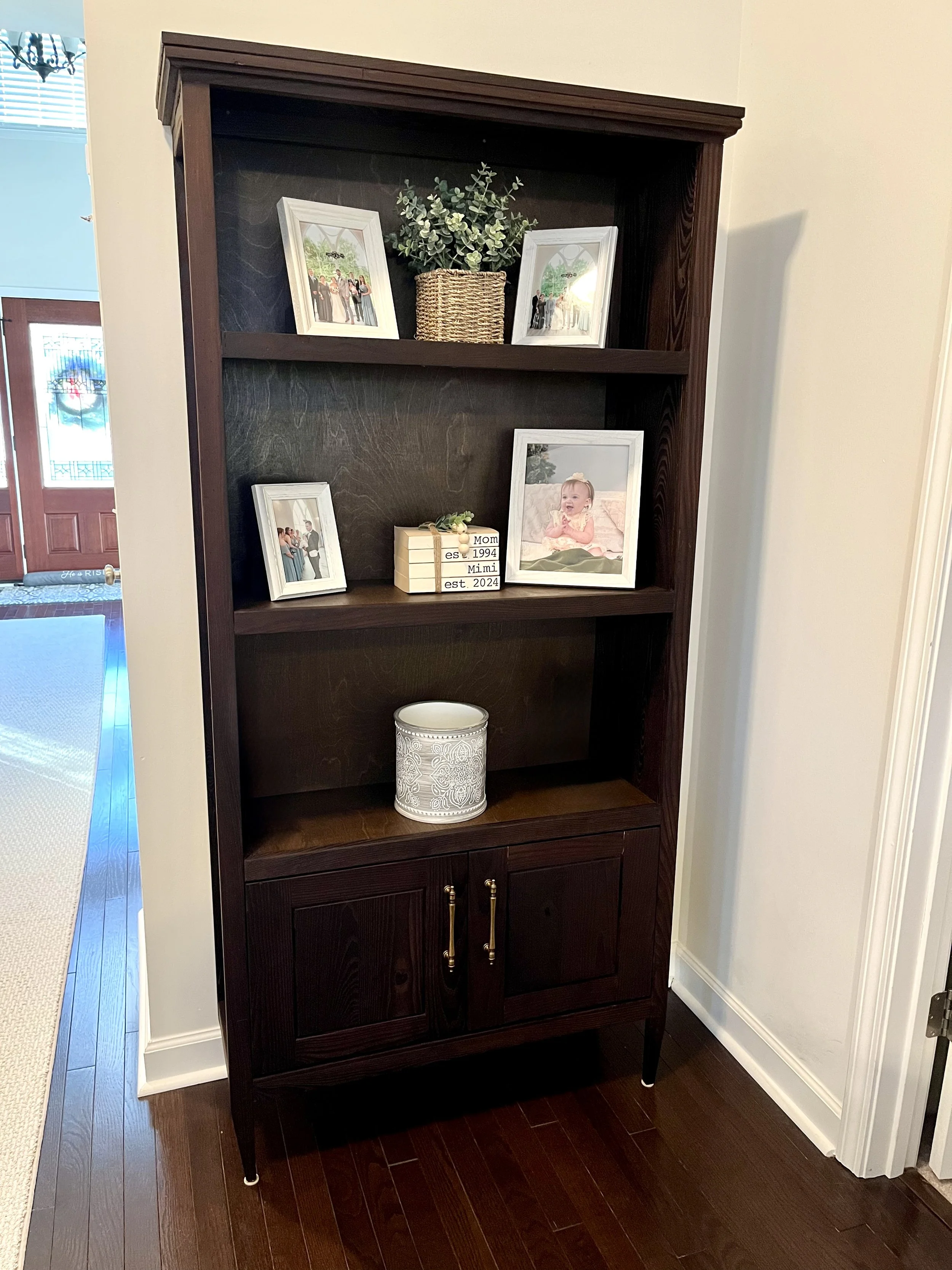 A dark wooden bookshelf with a top shelf holding framed family photos and a potted plant, and a middle shelf with a framed photograph and a decorative box with a note, in a room with hardwood floors and a white wall.