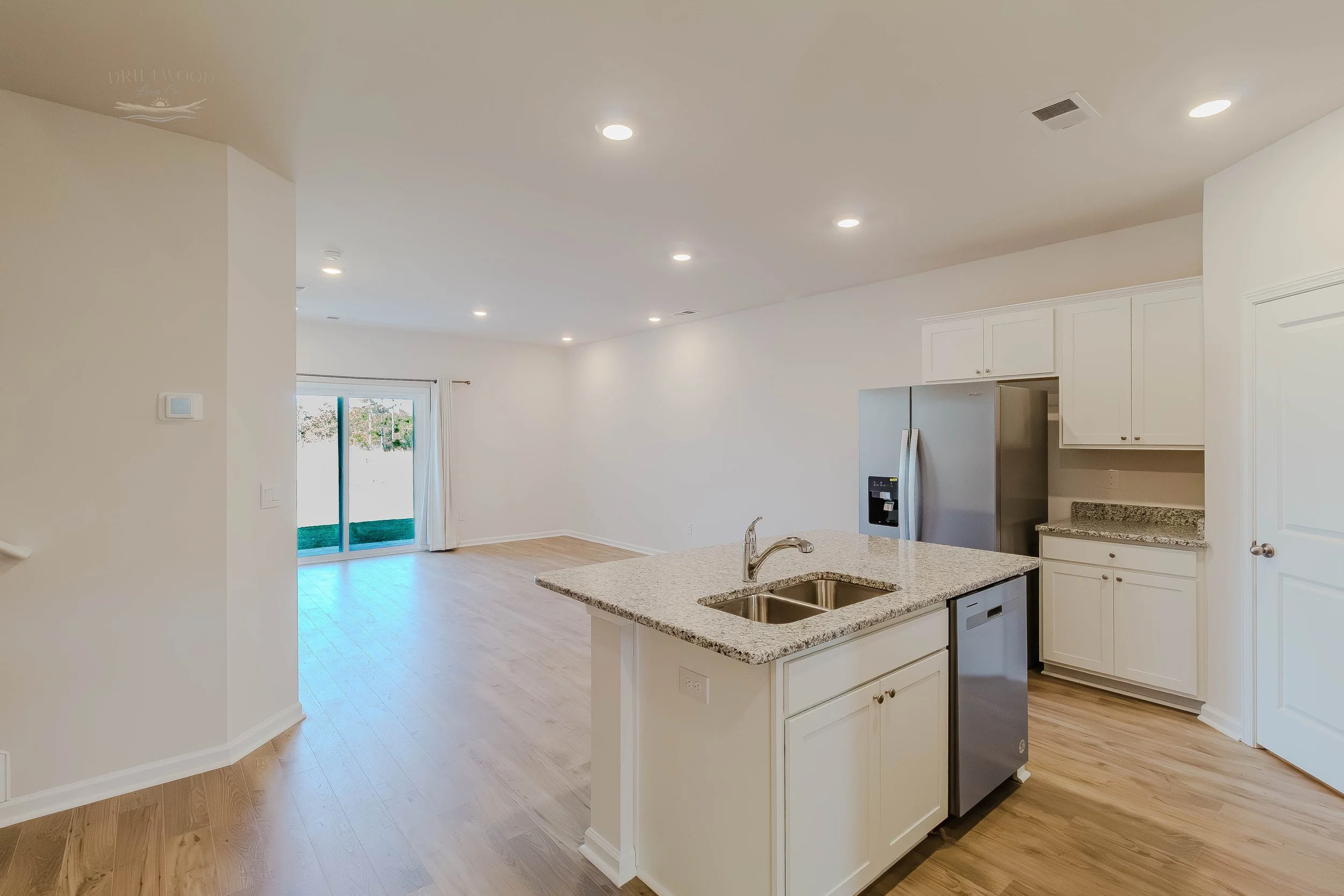 Modern open-concept kitchen with granite island, stainless steel refrigerator, white cabinets, and hardwood floors in a spacious living area with sliding glass door to the backyard.