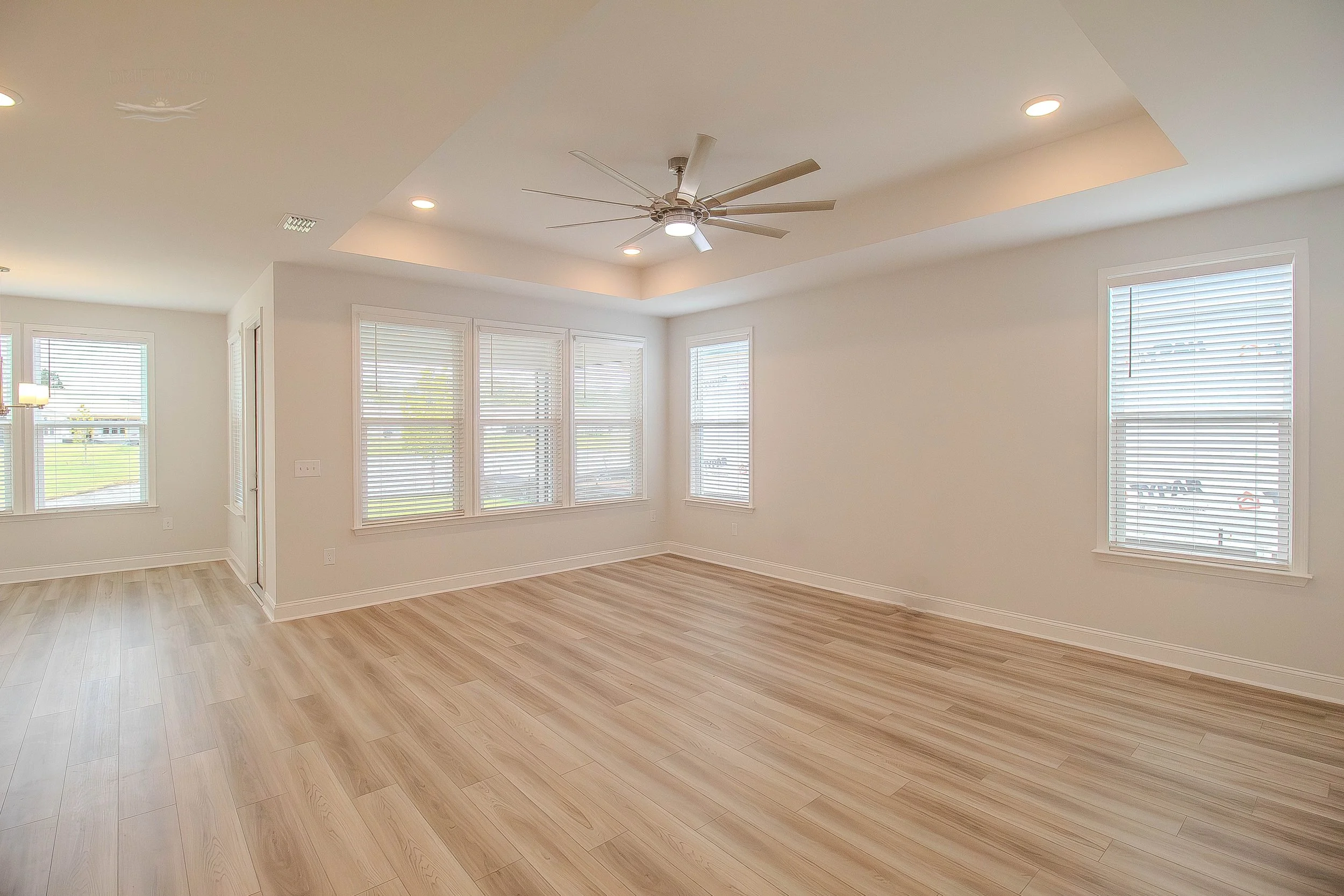 Empty living room with light wood floors, white walls, multiple windows with blinds, ceiling fan, and recessed lighting.
