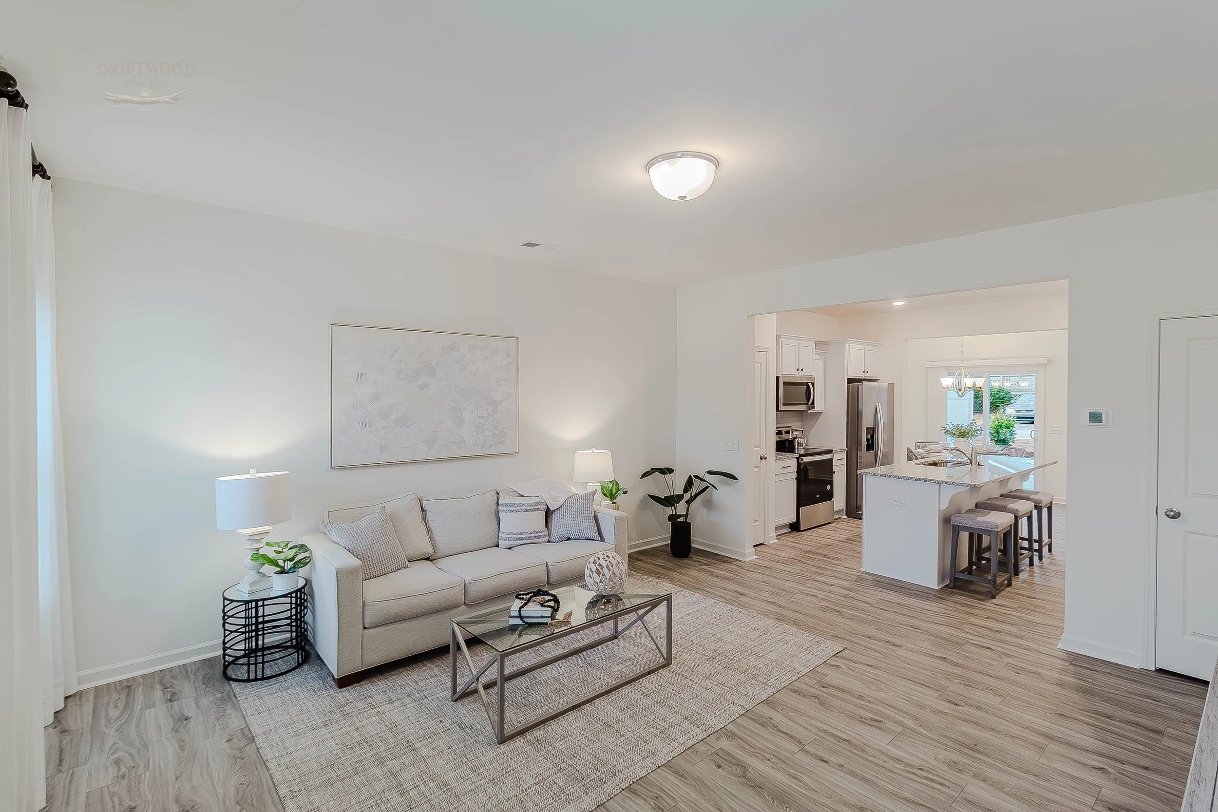A bright living room with a white sofa, side tables with lamps, and artwork on the wall, open to a kitchen with white cabinets, stainless steel appliances, and a breakfast bar with three stools, in a modern home.