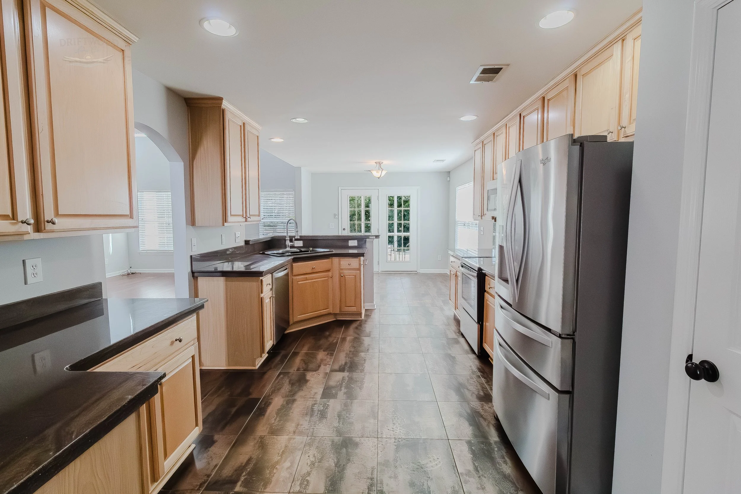 Modern kitchen with wooden cabinets, black countertops, stainless steel appliances, tile flooring, and a glass door leading outside.