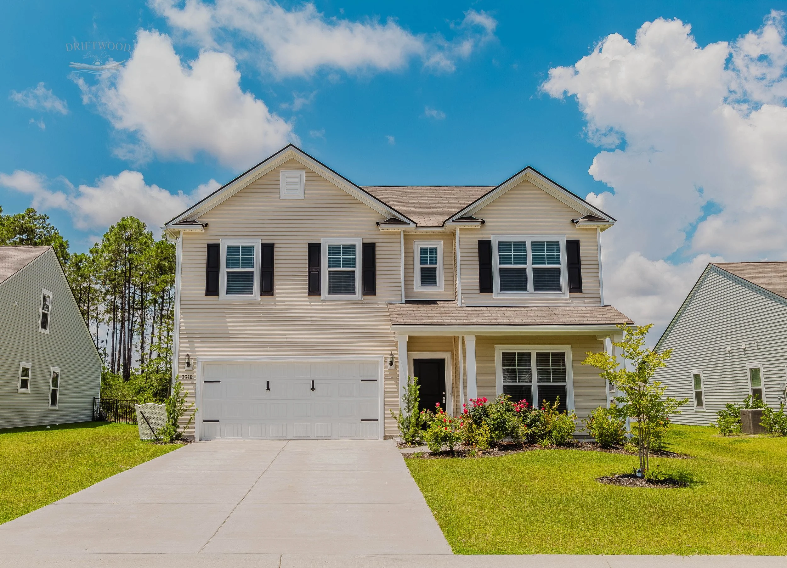 Modern two-story house with a white garage door, beige siding, black shutters, and a small front porch with plants, under a partly cloudy sky.