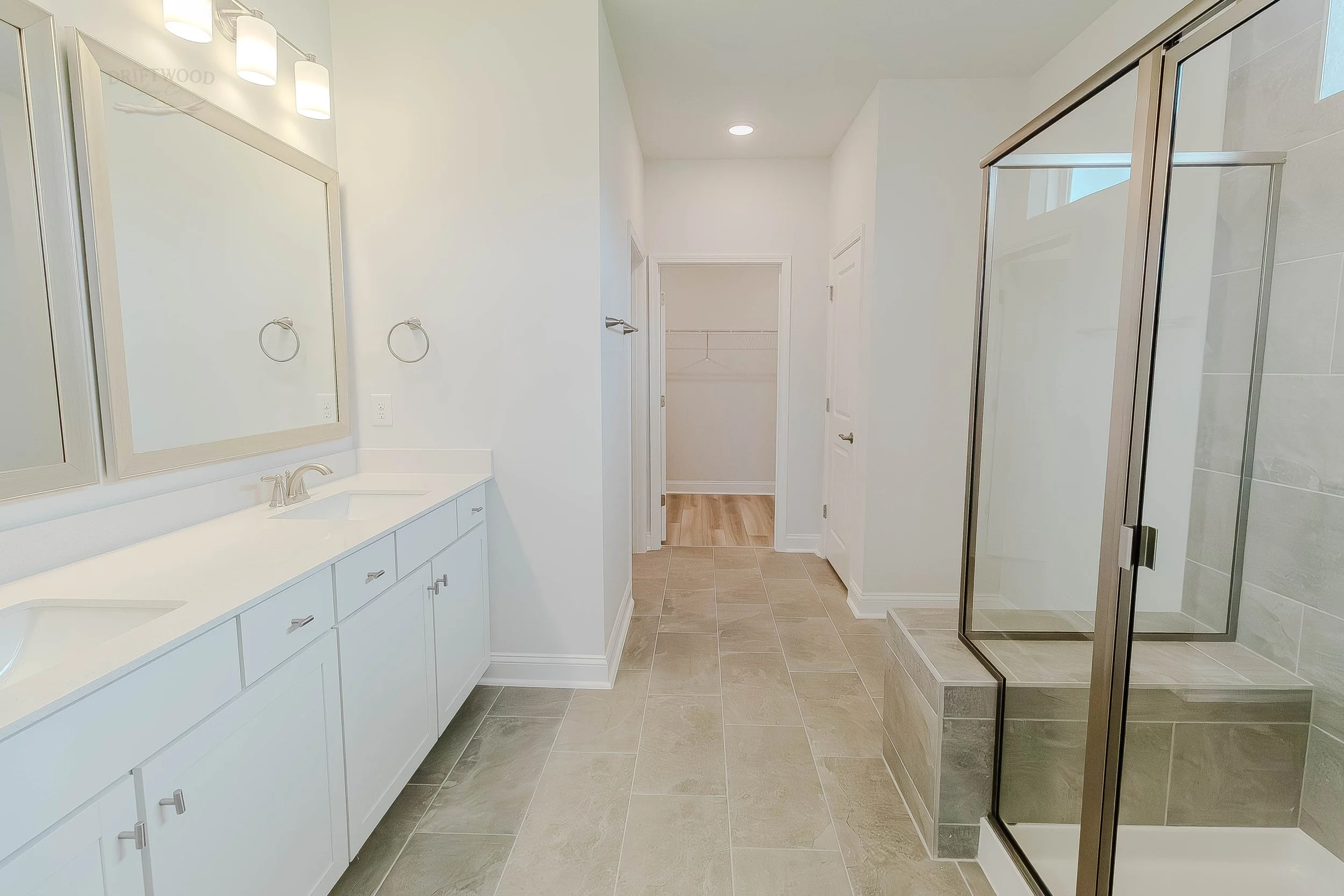 A spacious bathroom with a double sink vanity, large mirror, wall-mounted light fixtures, beige tiled floor, walk-in shower with glass door, and a walk-in closet at the end of the room.