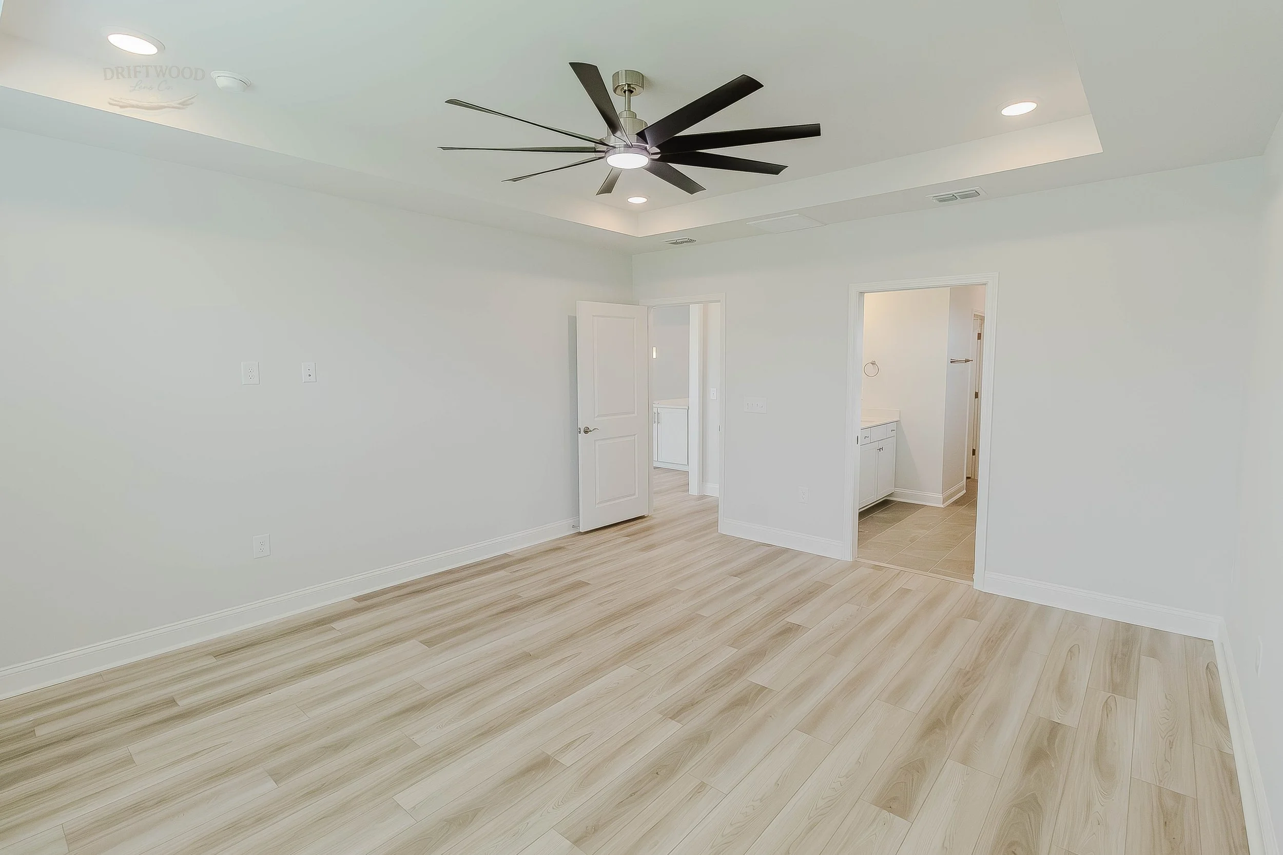 Empty room with white walls, light wood flooring, a ceiling fan, and two doorways leading to separate spaces.