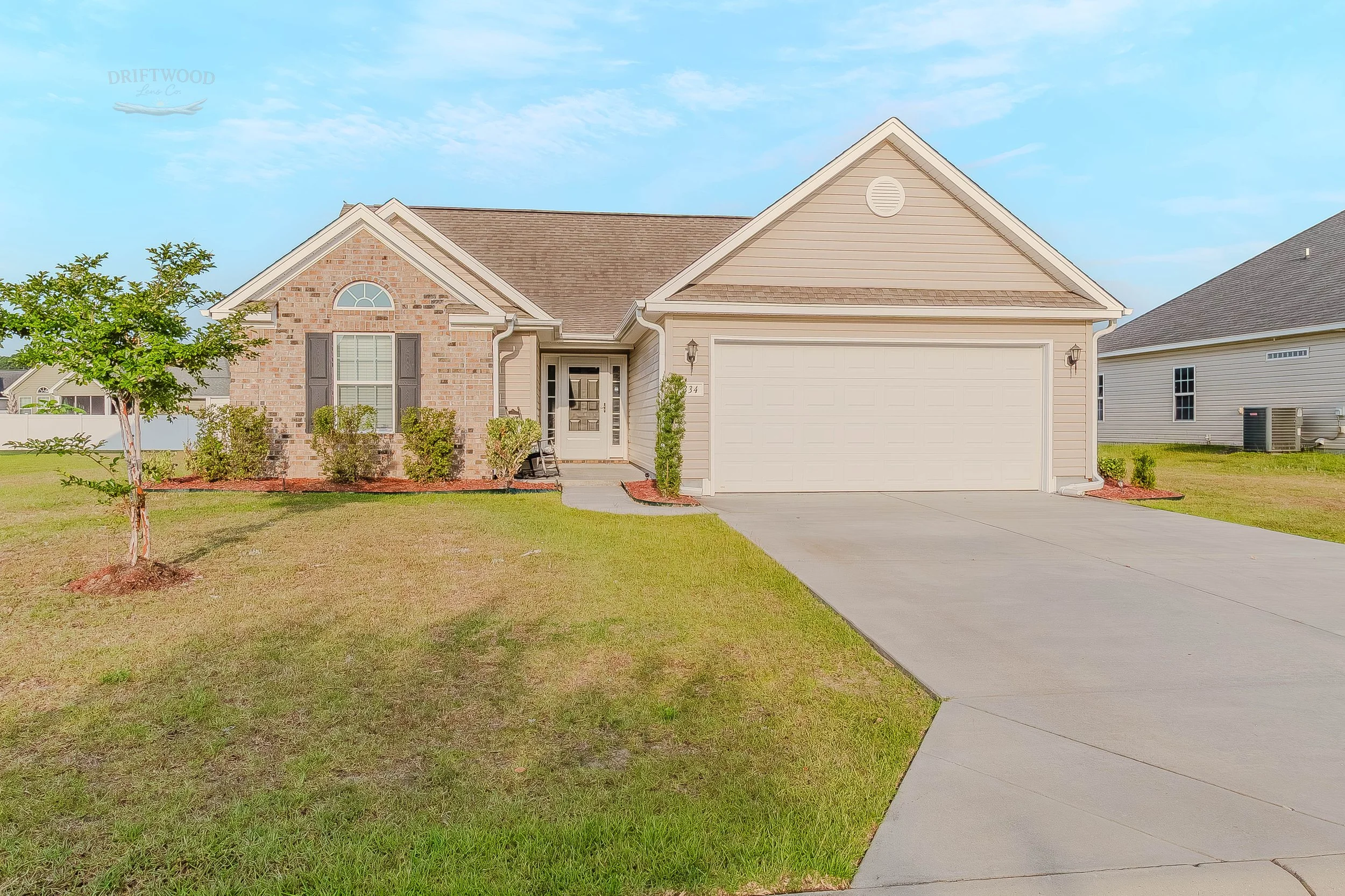 Front view of a suburban house with a brick and siding exterior, a two-car garage, and a well-maintained lawn with small trees and shrubs.