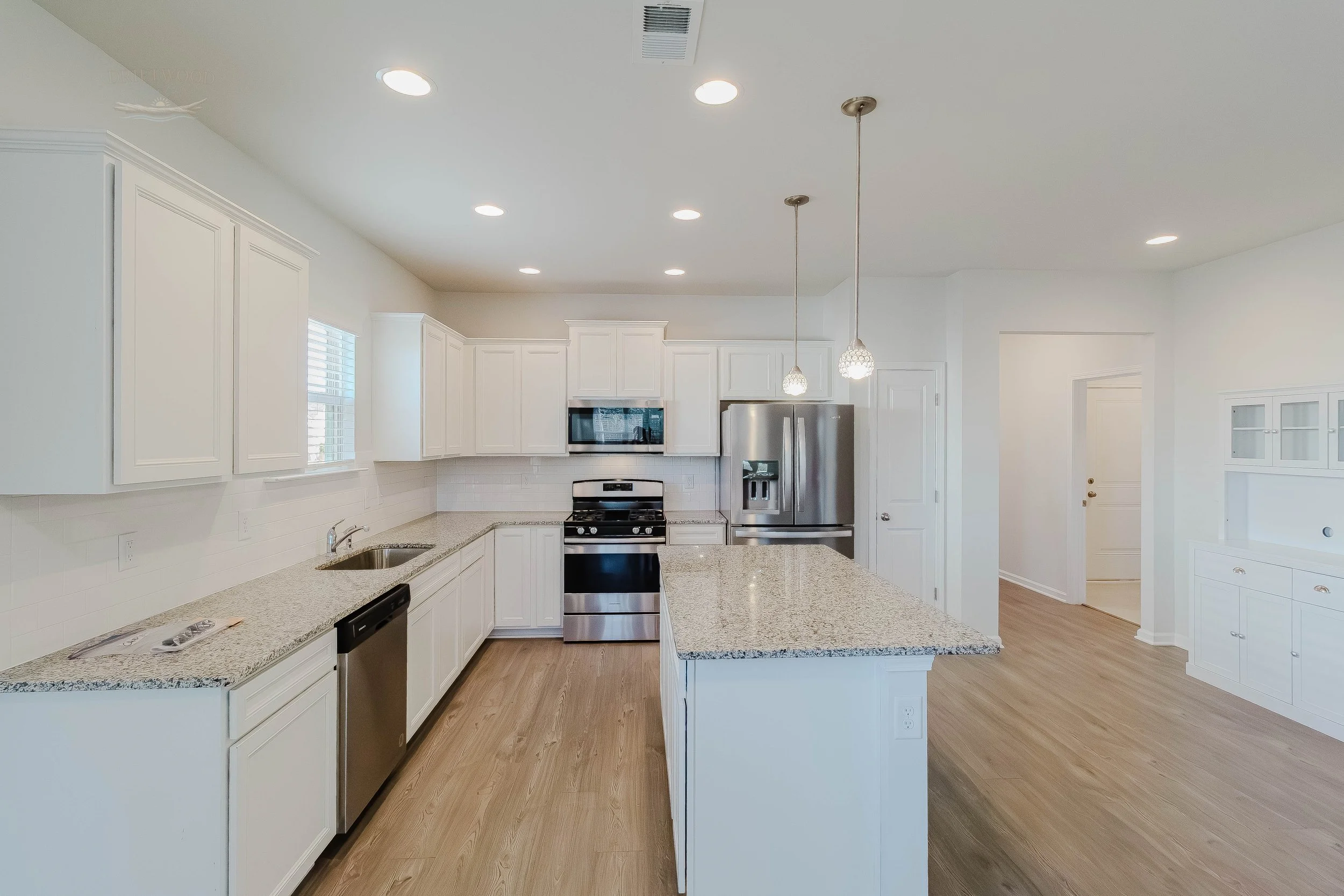 Empty modern kitchen with white cabinets, granite countertops, stainless steel appliances, and light wood flooring.