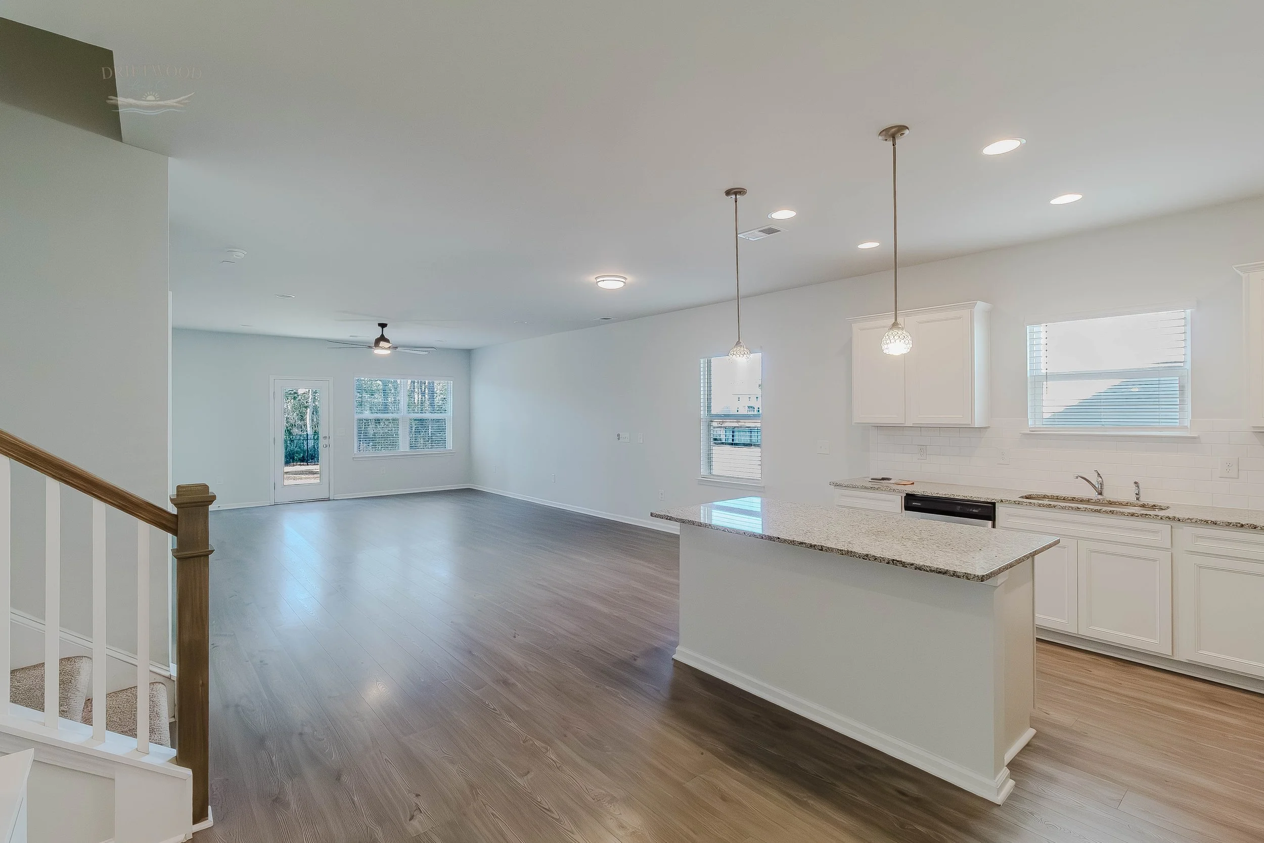 Open living area and kitchen with white cabinets, granite countertops, hardwood floors, pendant lights, large windows, and a door leading outside.