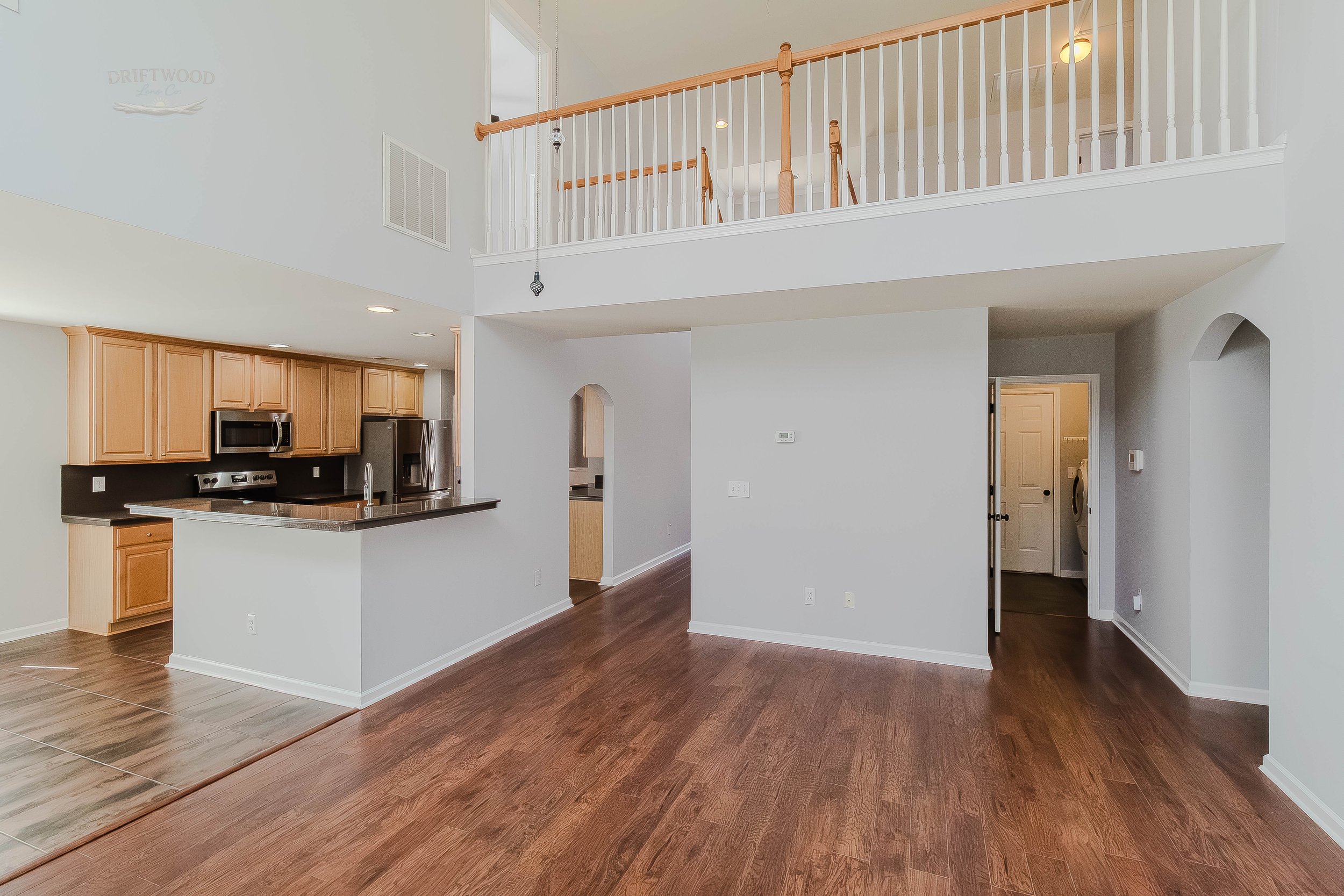 Open-concept living space with kitchen, hardwood floors, and a two-story ceiling. The kitchen has light wood cabinets, black countertops, and stainless steel appliances. There is a balcony or upper-level railing visible from the living area.