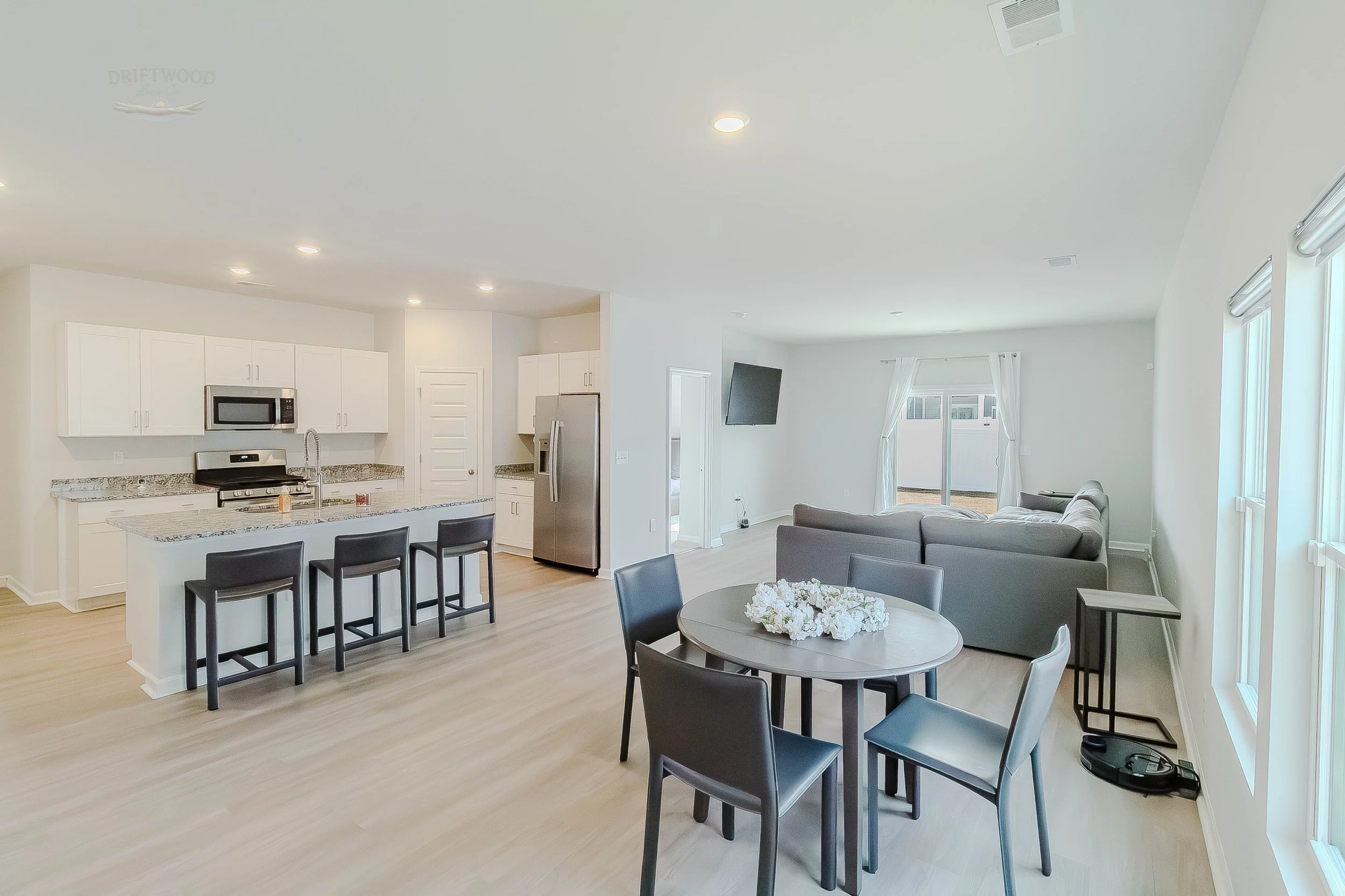Open-concept living room and kitchen area with white cabinetry, stainless steel appliances, a granite countertop island with three black chairs, a round dining table with five black chairs, a gray sectional sofa, and windows letting in natural light.