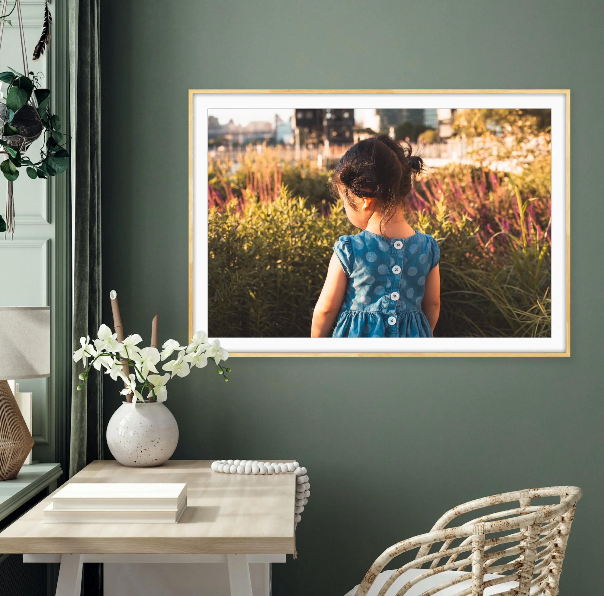 Interior of a room with a green wall, a framed picture of a girl facing away outdoors, a white table with books, a large white vase with white orchids, a beaded decoration, and rattan chairs.