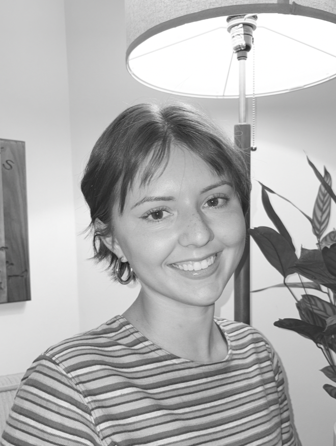 A young woman with short hair smiling, wearing a striped shirt, standing next to a tall floor lamp and near a potted plant.