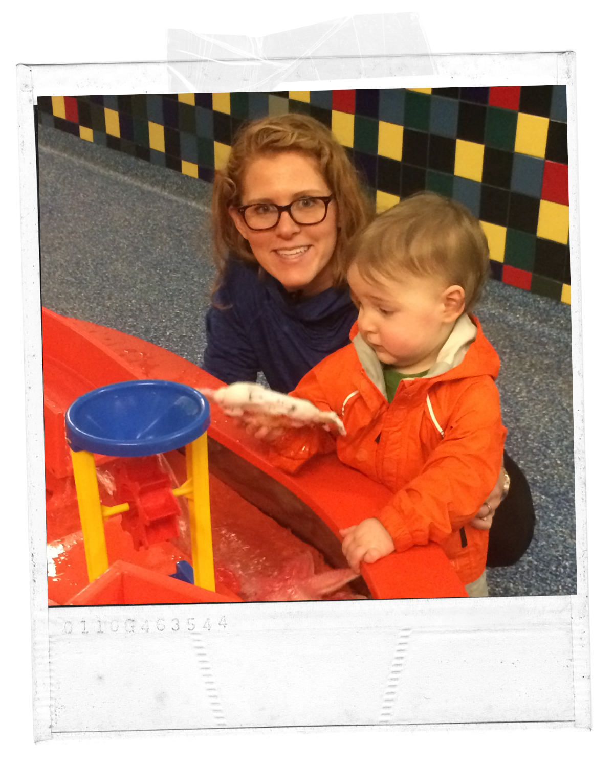 A woman with glasses and curly red hair smiling at a young boy with blond hair, who is looking at a device in his hand. They are at an indoor play area with bright, colorful tiled walls and a red table.