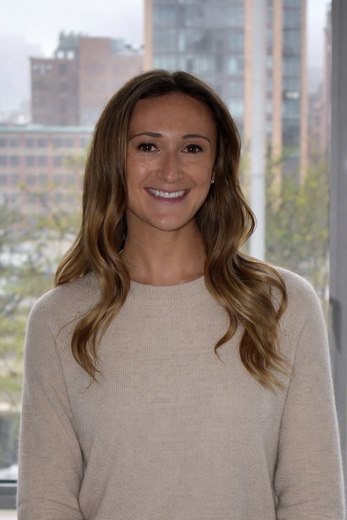 Smiling woman with wavy light brown hair wearing a beige sweater, standing indoors with a cityscape background visible through a large window.