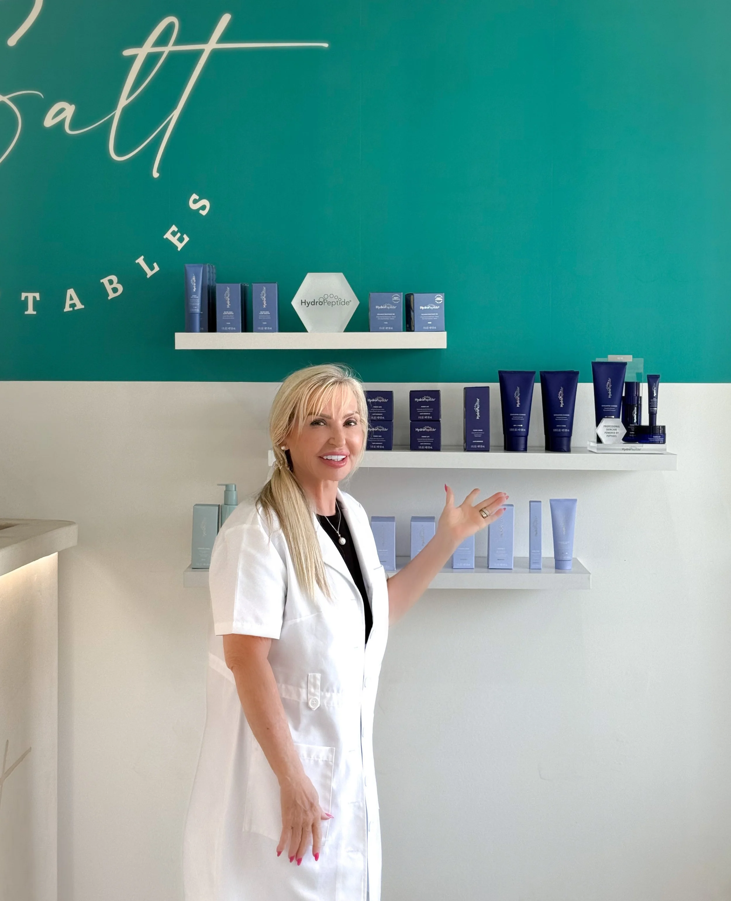 A woman in a white lab coat standing in front of shelves with skincare products, smiling and gesturing towards the products, with a teal wall behind her that says 'Salt' and 'Tablets'.