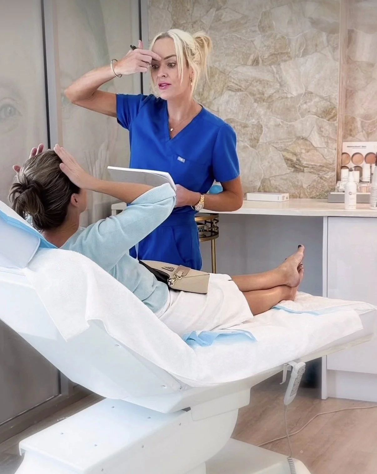 A woman in blue medical scrubs is talking to a patient lying on an examination table in a medical office or clinic.