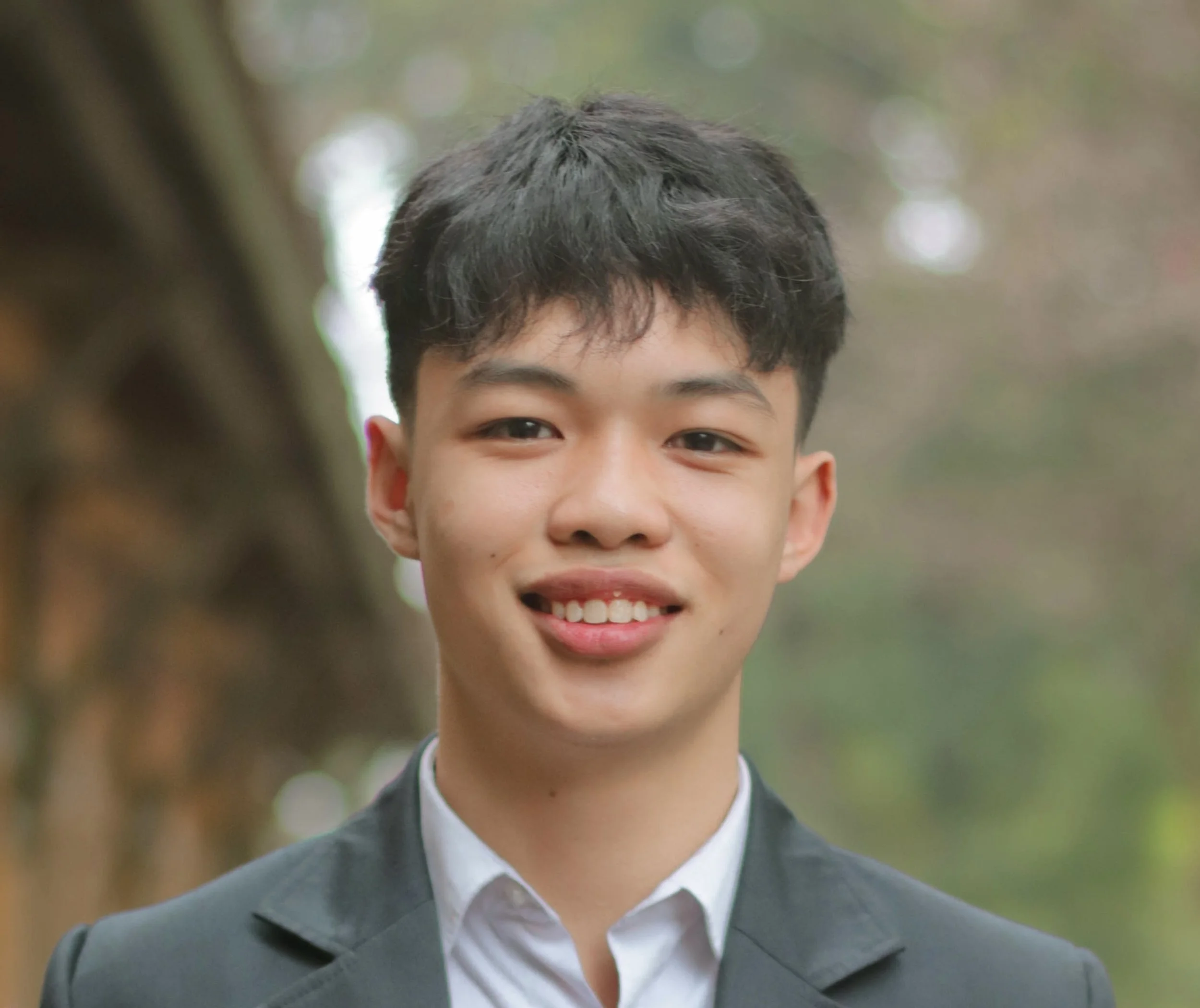 Close-up of a young person with short dark hair wearing a suit and tie, smiling outdoors with blurred greenery in the background.