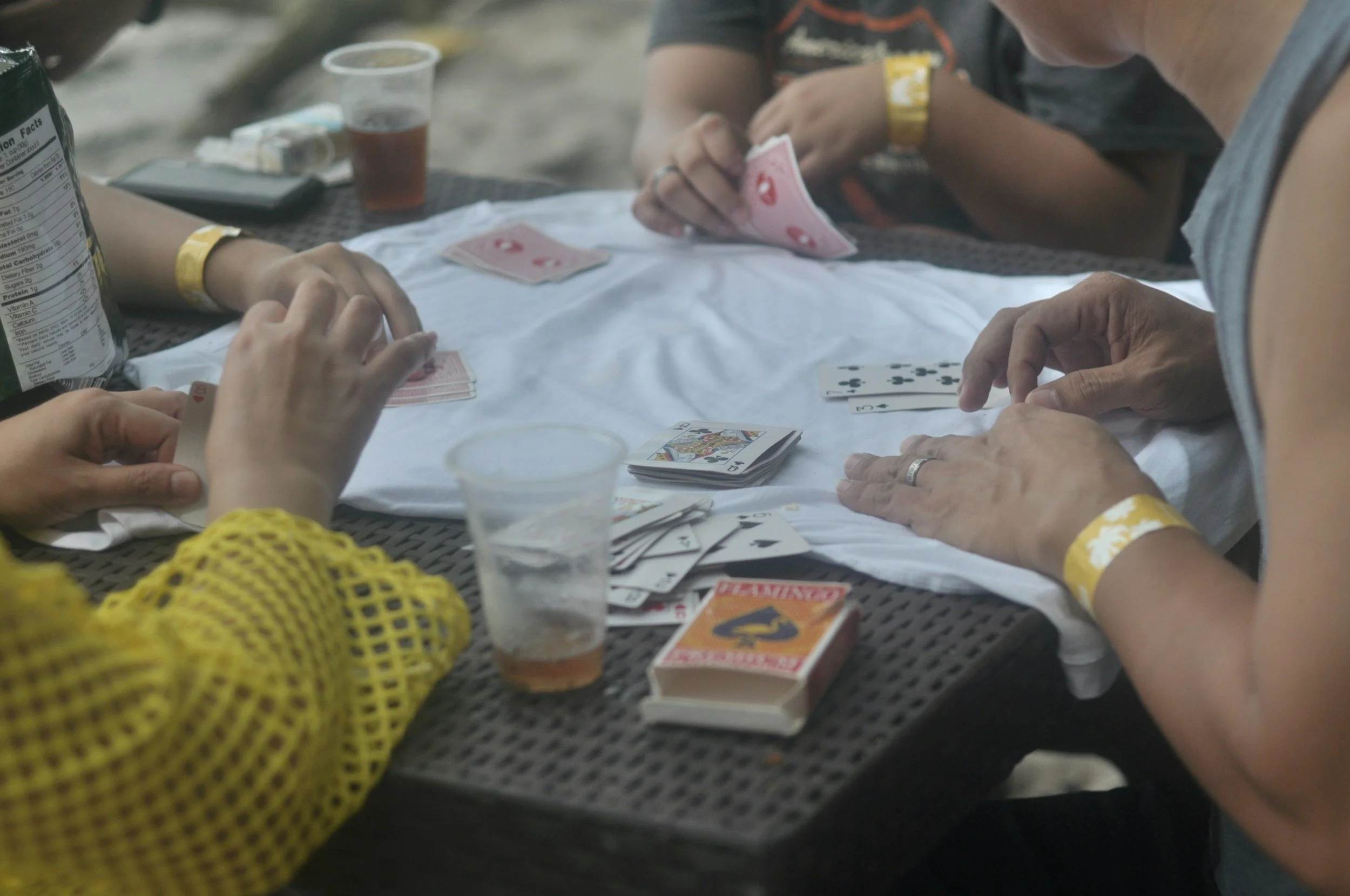 People playing a card game, likely hearts, at an outdoor table with drinks and snacks, one person wearing a yellow crocheted sweater and another with a gray sleeveless shirt.