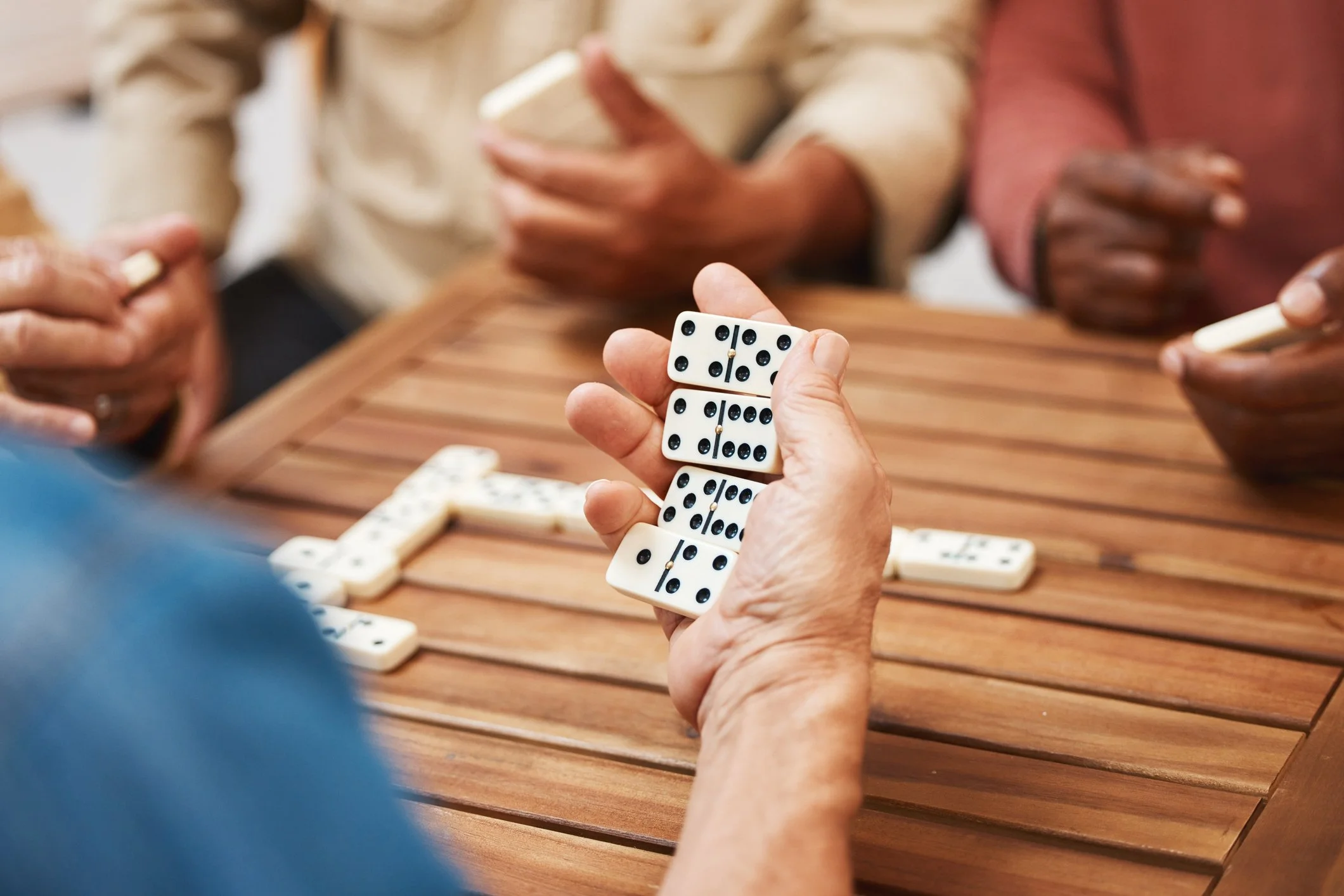 Close-up of a person holding dominoes in front of a group of people playing dominoes at a wooden table.