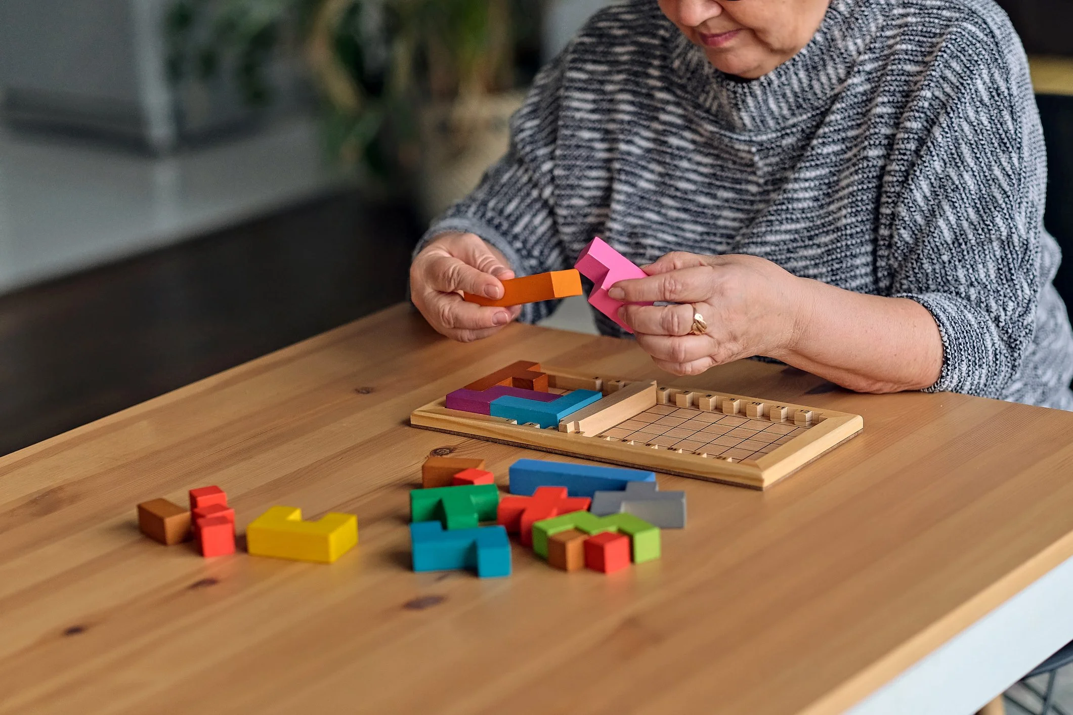 An woman wearing a gray and black striped sweater is playing a colorful board game with various small, brightly colored wooden blocks on a wooden table.