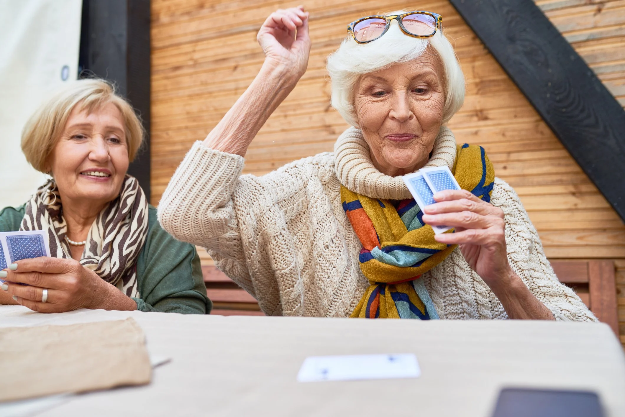 Two elderly women playing cards at a table outdoors, one adjusting her glasses while the other smiles.