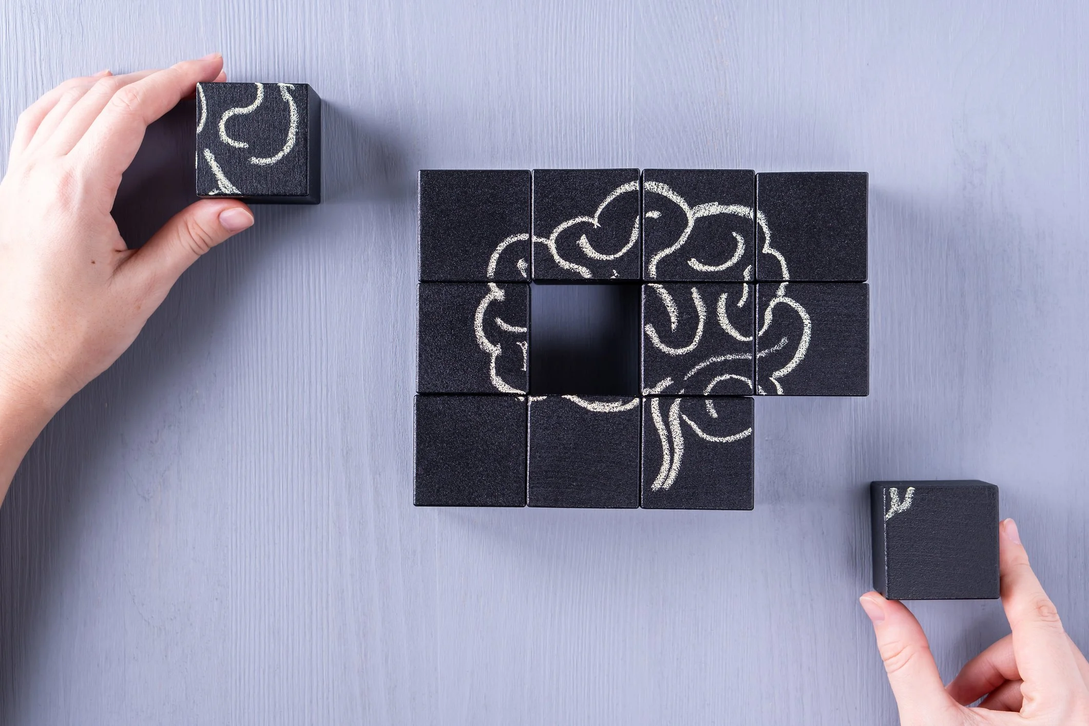Person's hands arranging black blocks with white chalk drawings of a brain on a light gray surface.