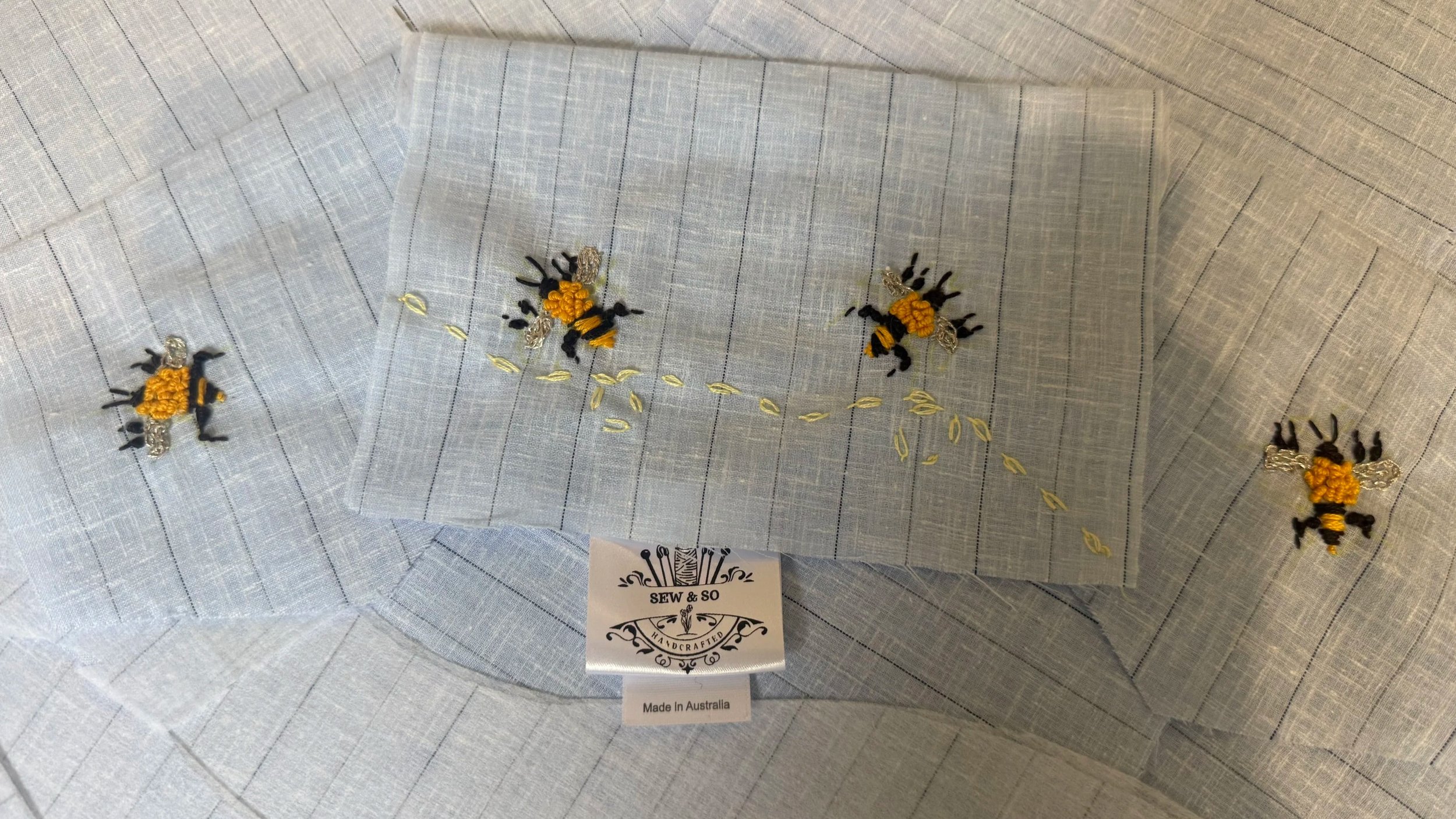 A light blue piece of fabric embroidered with honeybee designs and yellow pollen. The embroidery features four bees and a trail of pollen grains, with some nearby embroidered leaves. The fabric tag indicates it is handmade in Australia and from SEW & SO.