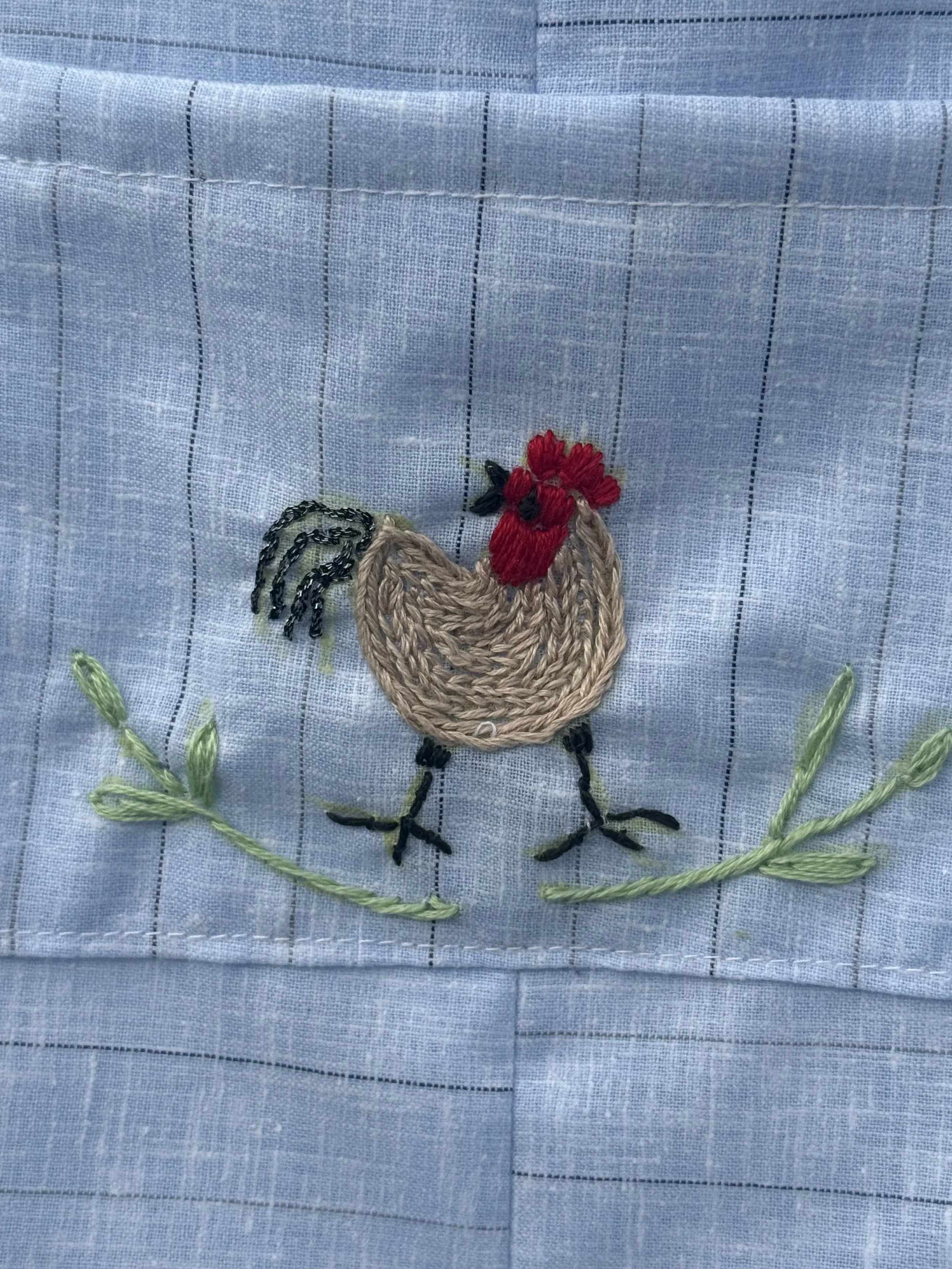 Embroidery of a chicken with a red comb, black beak, and black legs on light blue fabric with green embroidered grass.