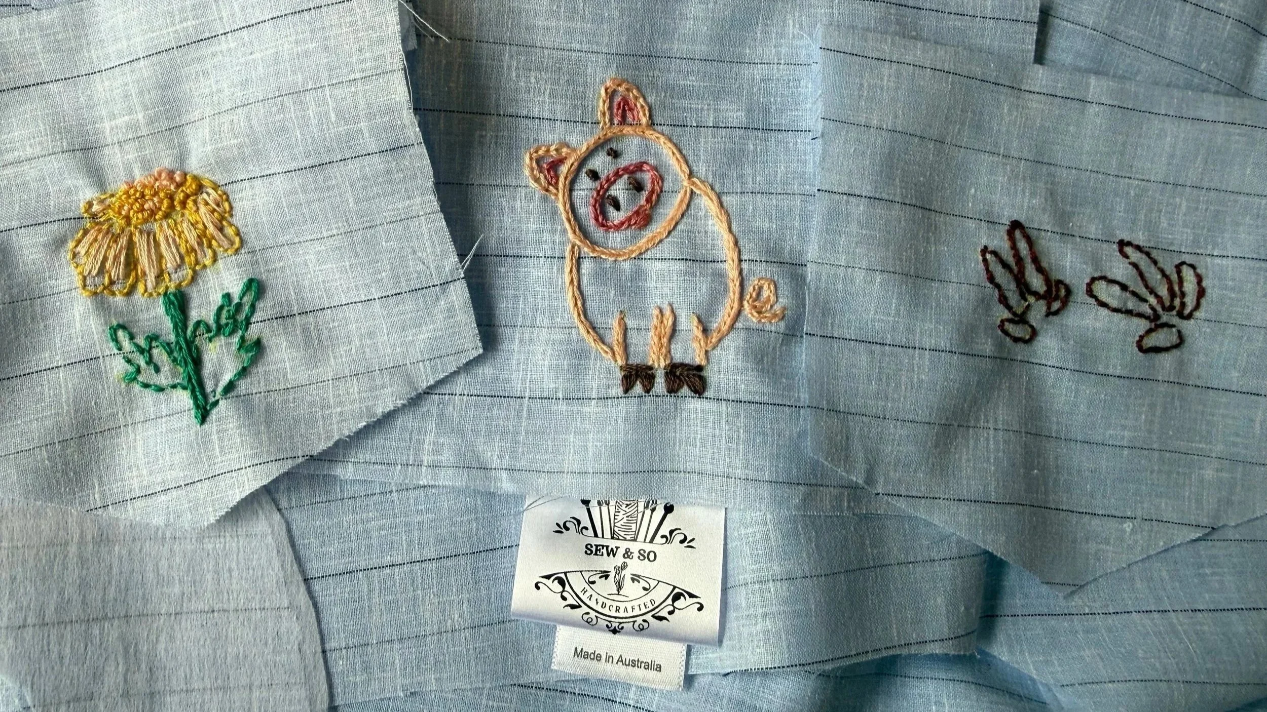 Light blue fabric with three embroidered patches: a yellow flower with green stem and leaves on the left, a pink pig with black eyes and hooves in the center, and two small black embroidered bunny ears on the right. There is a white tag underneath that reads 'SEW & SO HANDCRAFTED Made in Australia'.