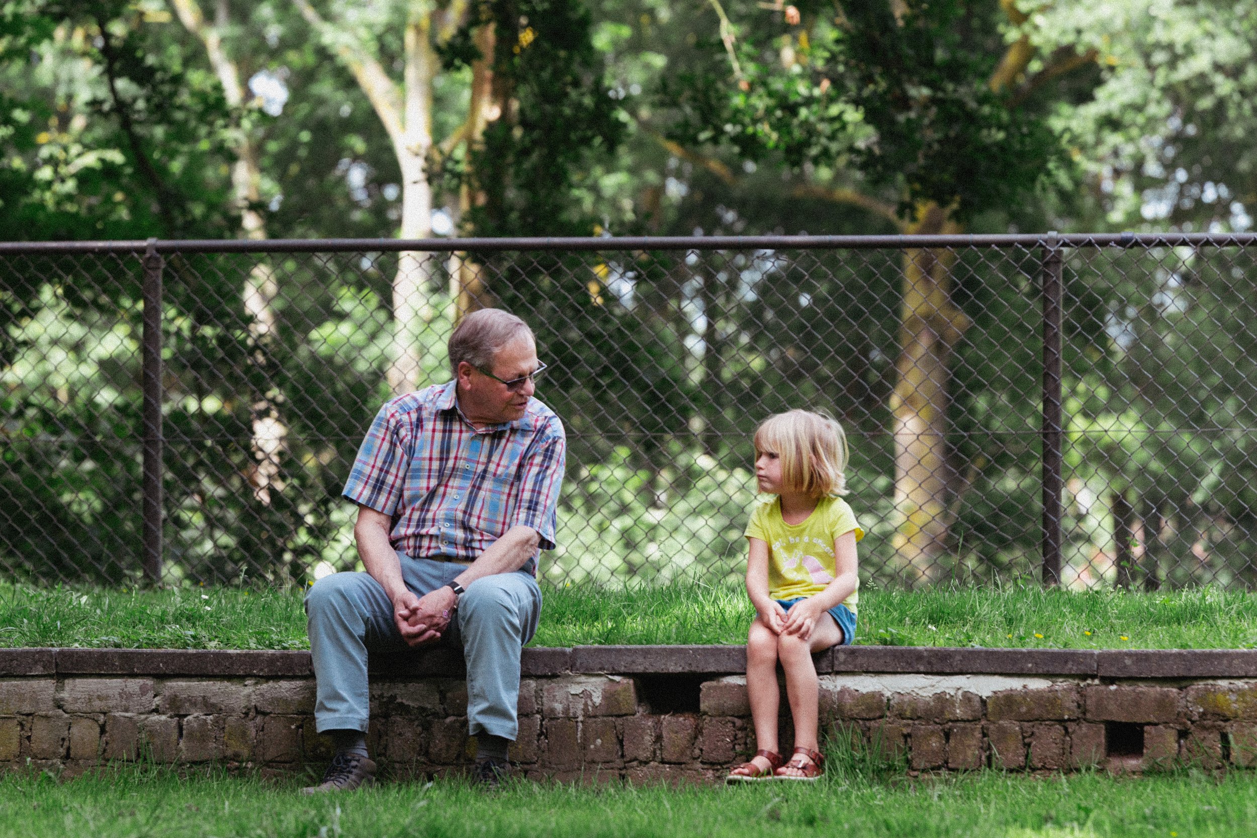 An elderly man and a young girl sitting on a park bench in front of a black wire fence, surrounded by green trees.