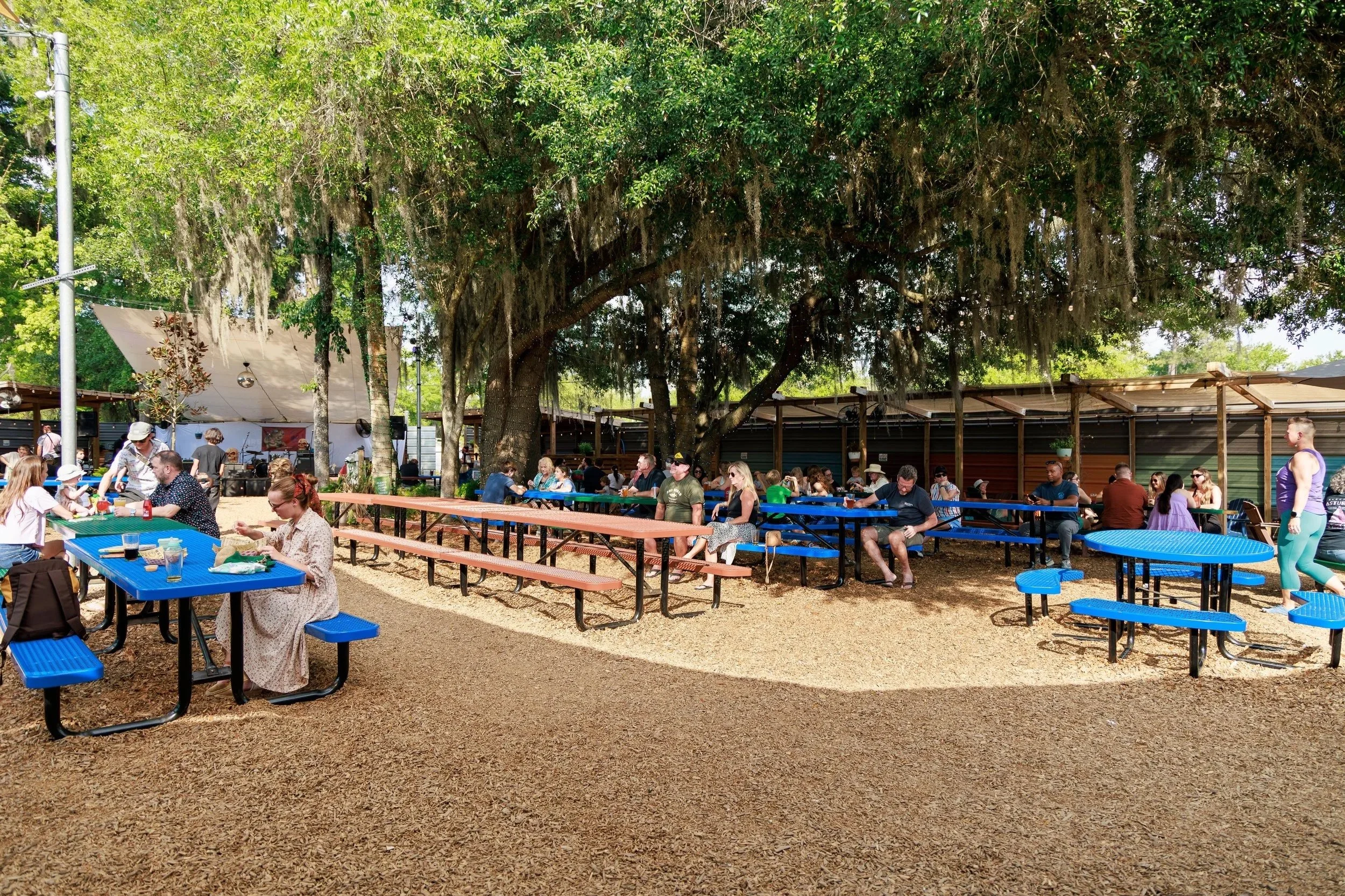People sitting at colorful picnic tables under a large tree in a park, with some standing and socializing, and a stage with sound equipment in the background.