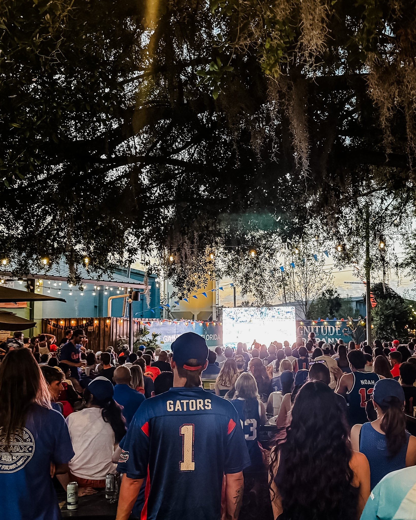 Crowd of people watching an outdoor concert, with a large screen displaying the band 'Gritty' and festive string lights hanging overhead. Some attendees are wearing sports jerseys, including a University of Florida Gators jersey.