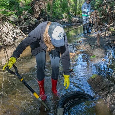 Two people are working in a shallow stream in a wooded area, using tools like a long-handled device and a coil, with one person wearing yellow gloves and red boots.