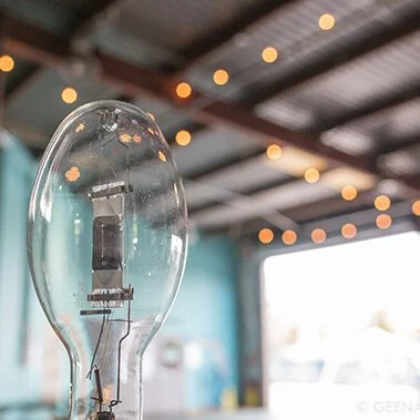 Close-up of an incandescent light bulb with visible filament, hanging in a decorated space with string lights and a roof structure in the background.