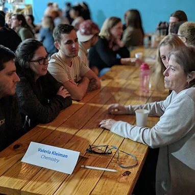 Group of students sitting at a wooden table, listening to a woman speaking, with a sign indicating 'Valeria Kleiman, Chemistry'.