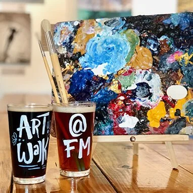 A painting palette with various colors of paint, two glasses labeled 'Art Walk' and '@ FM' with paintbrushes inside, on a wooden table.