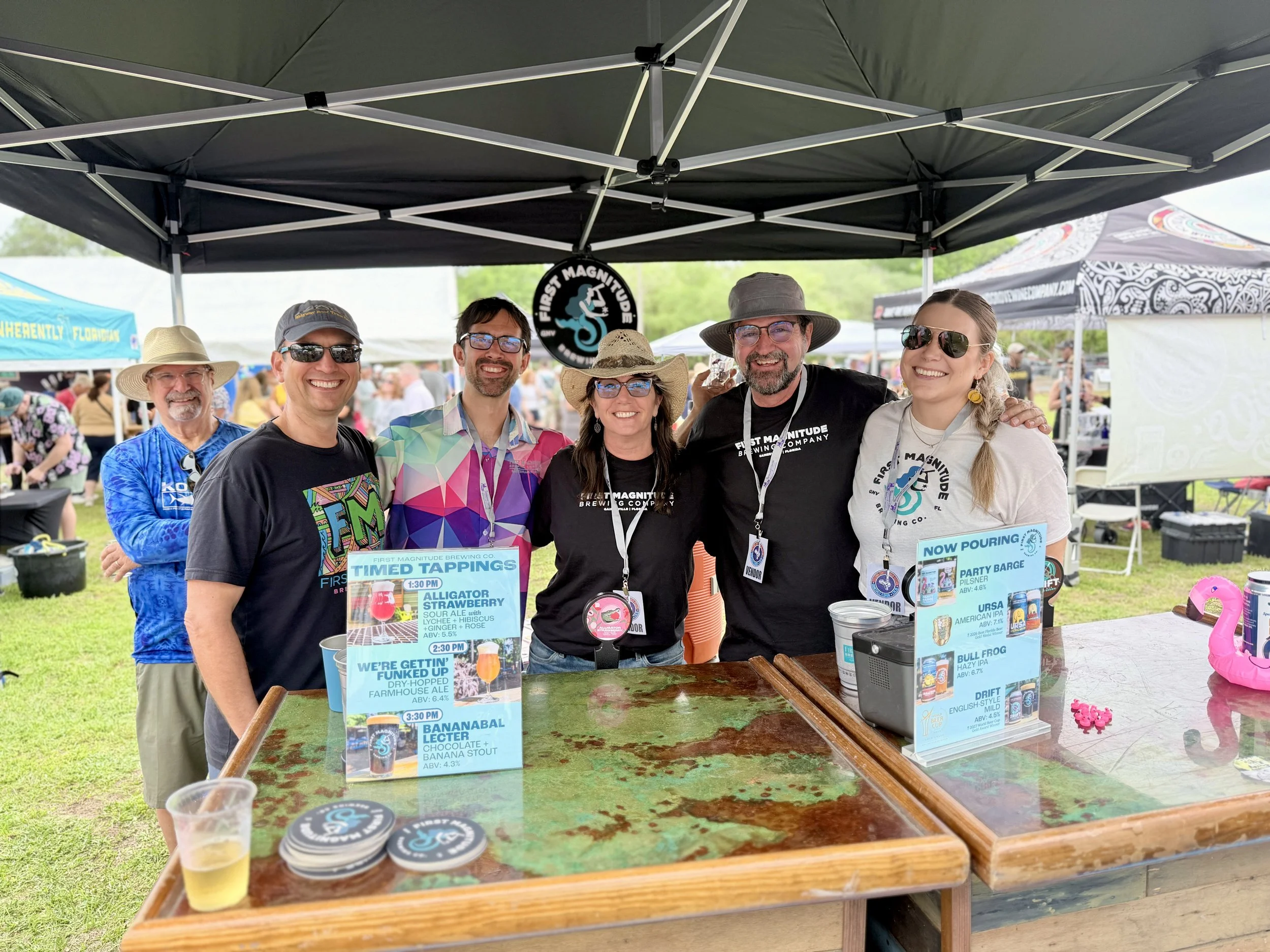 Group of six people standing under a black canopy tent at an outdoor event, smiling for the camera. The table in front displays beer tasting signs with different beer names and descriptions.