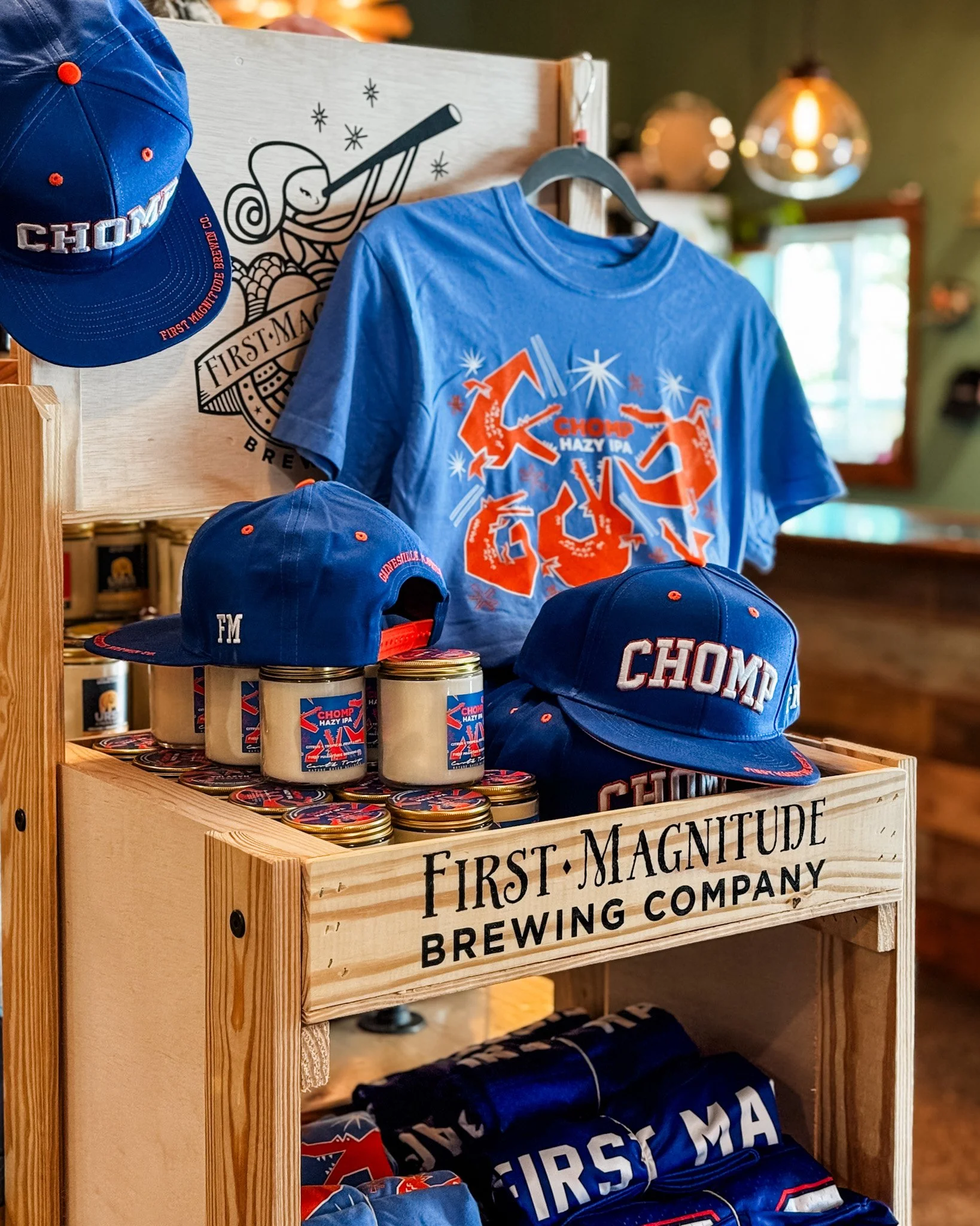 Display of apparel and merchandise from First Magnitude Brewing Company, including blue hats with red accents, blue T-shirts, and cans of beer, arranged on a wooden rack with the company's name printed on it.