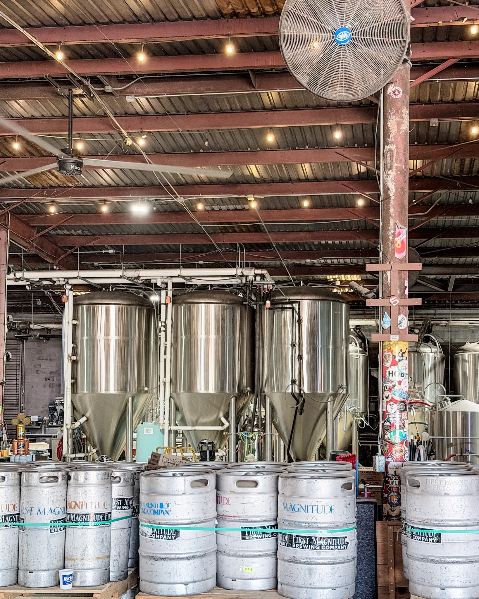 View of the interior of a brewery with large stainless steel fermentation tanks, beer kegs, and industrial equipment, with a ceiling fan and hanging lights.