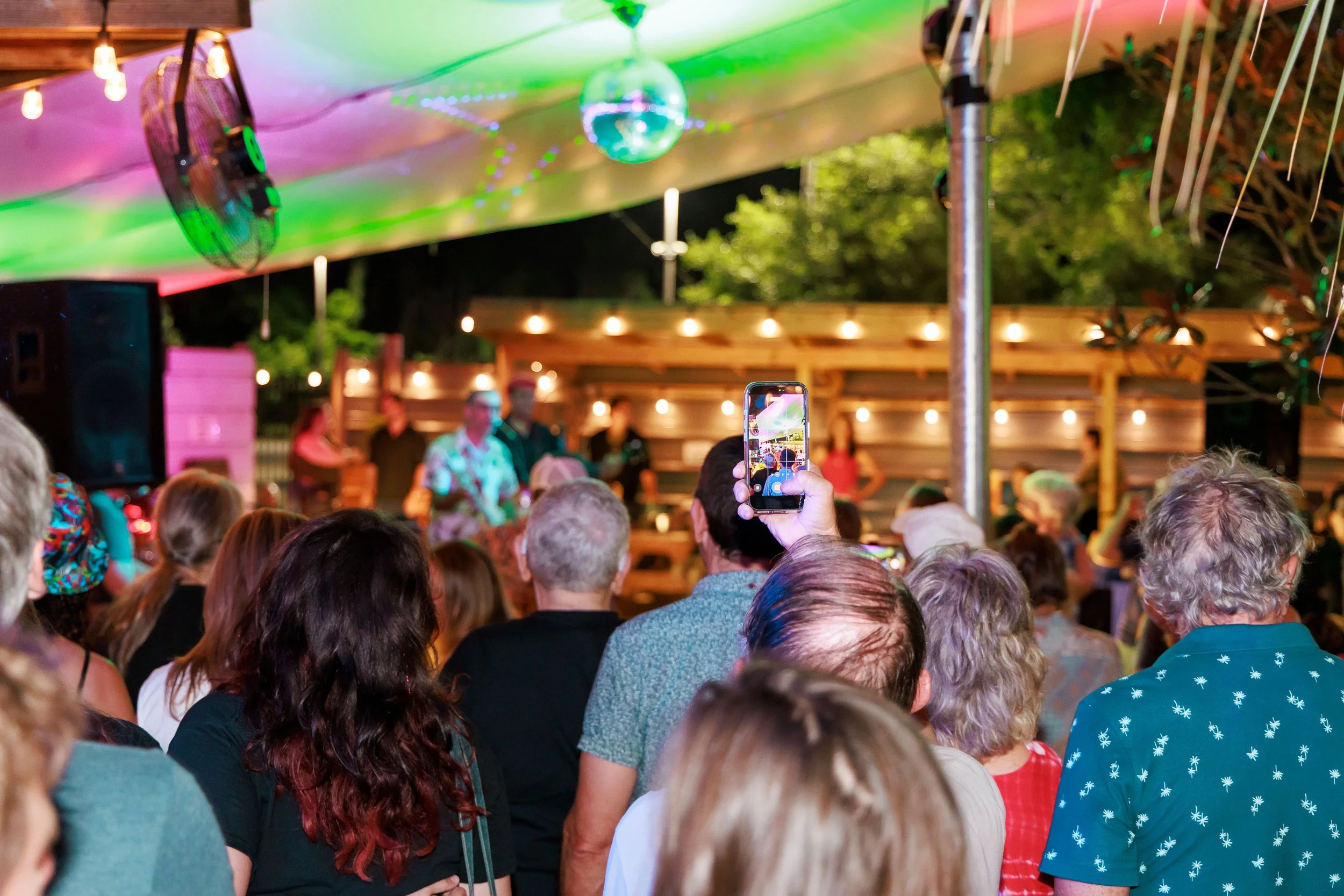 Crowd gathered at an outdoor event with a wooden stage, string lights, a disco ball, and colorful lighting. Some people are recording with their phones.