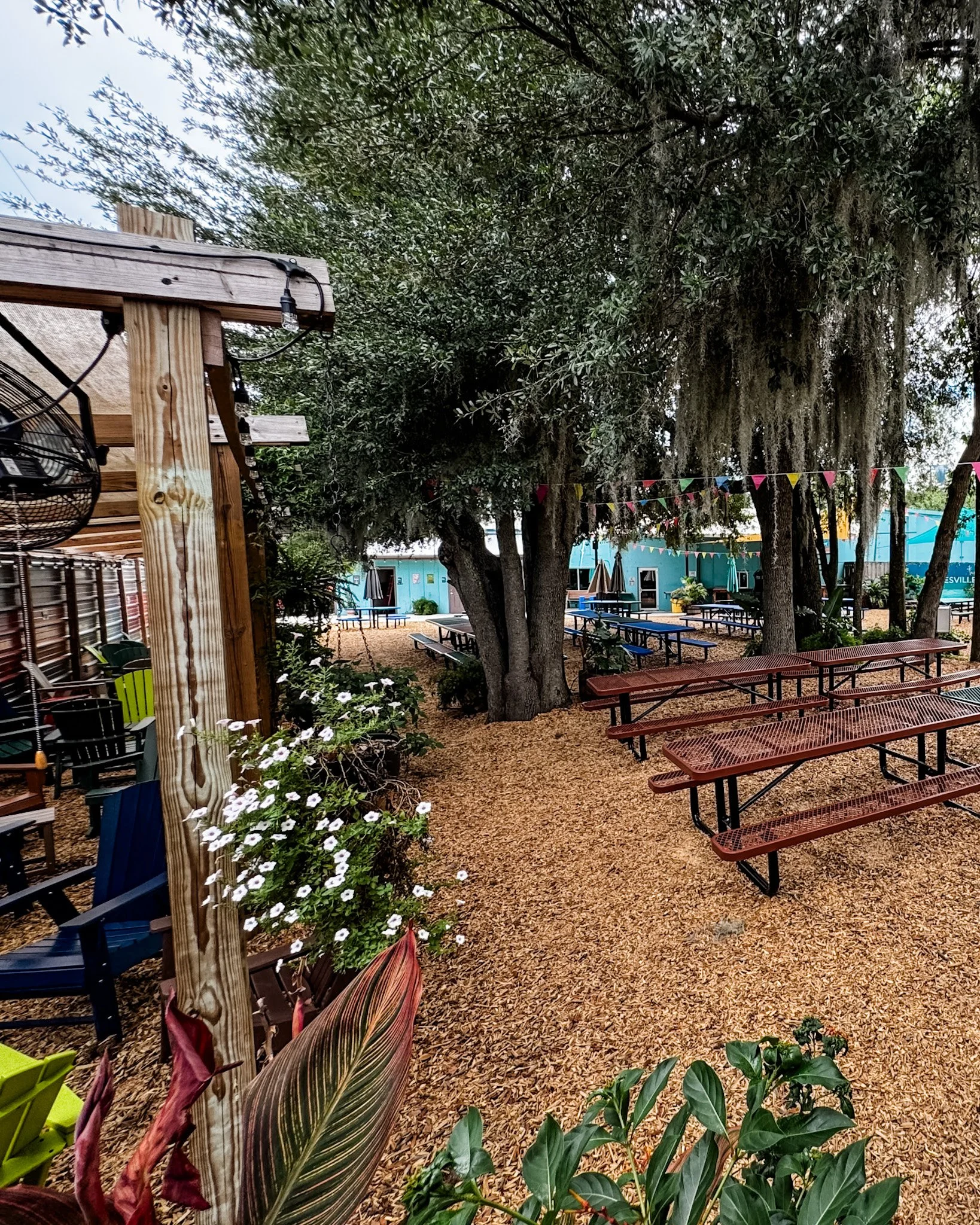 Outdoor seating area with wooden tables and benches, large trees, string lights and colorful bunting flags, with a blue building in the background.