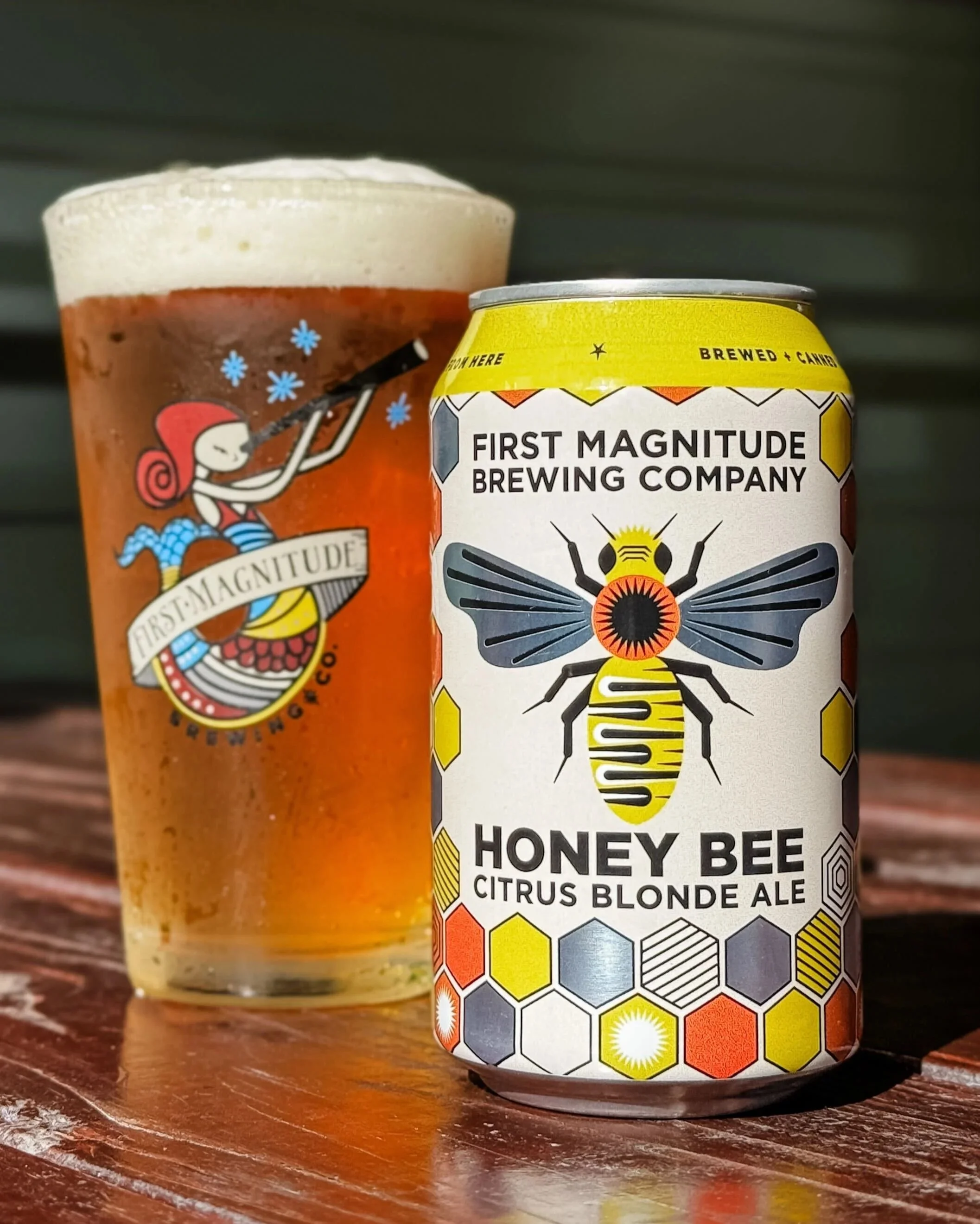 A can of First Magnitude Brewing Company Honey Bee Citrus Blonde Ale with a honey bee graphic, in front of a glass of beer on a wooden surface.