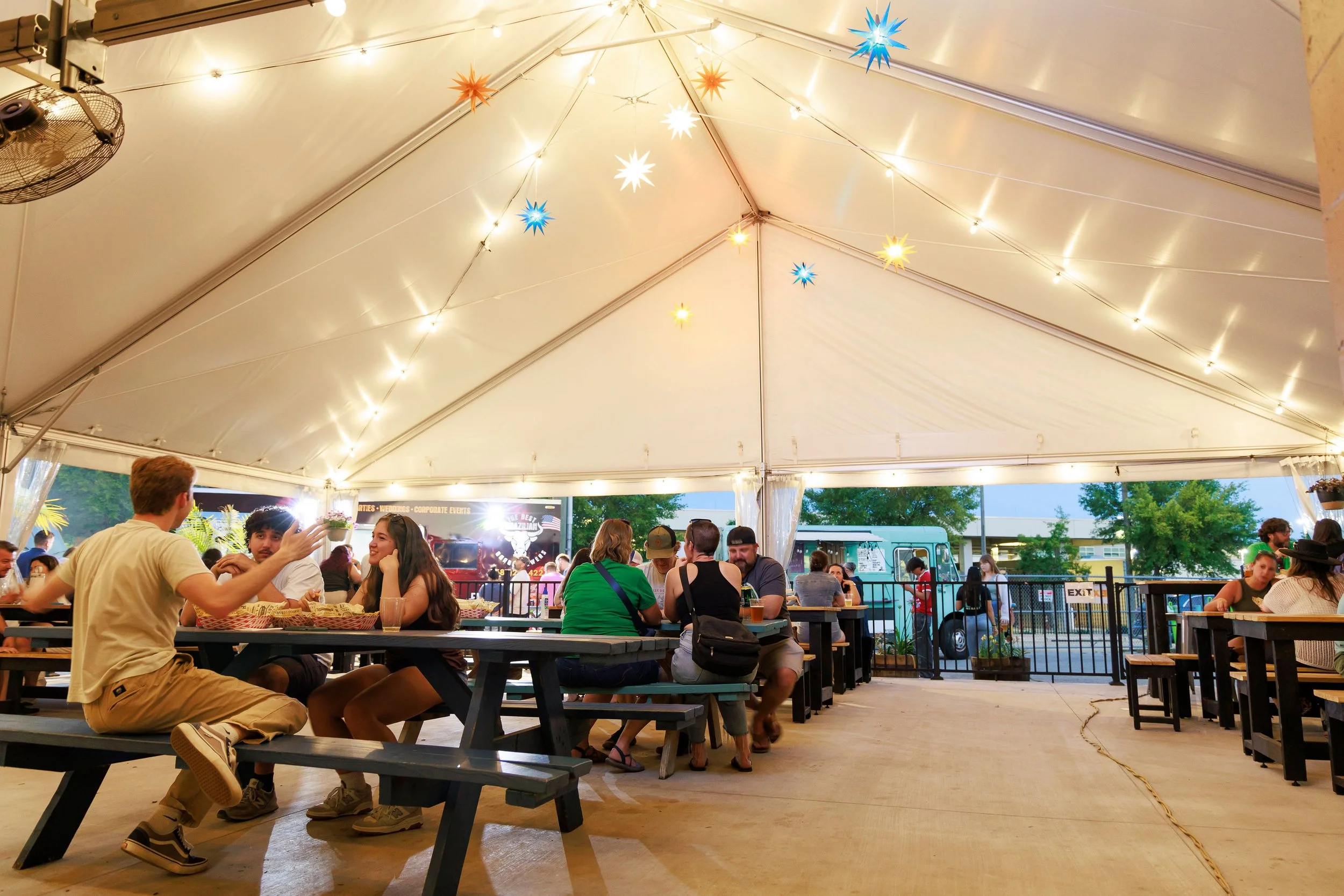 People sitting and socializing at picnic tables inside a large white tent with string lights and colorful star-shaped decorations hanging from the ceiling. The outdoor setting is visible through the open sides of the tent.