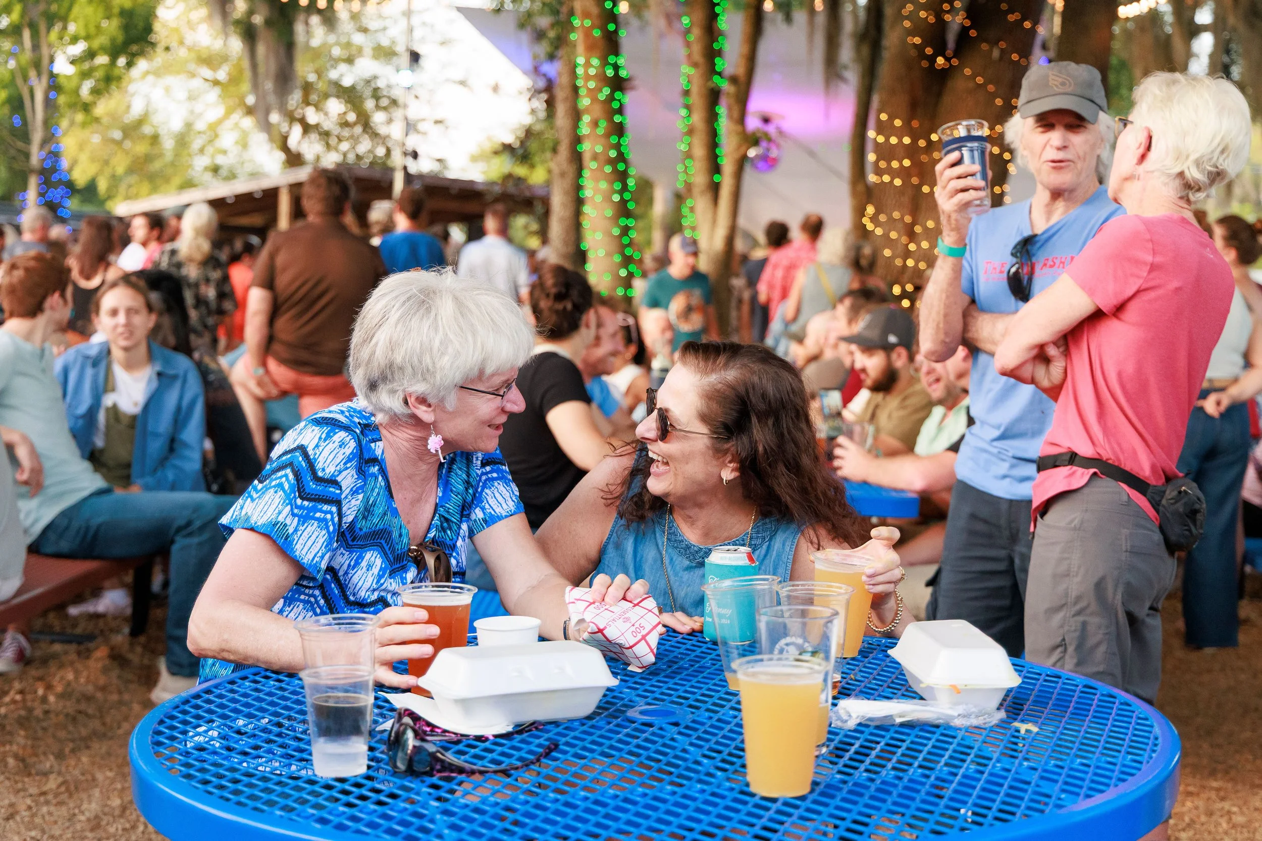 People socializing outdoors at a gathering in a wooded area decorated with string lights, with some sitting at a blue table enjoying drinks and others standing and chatting.
