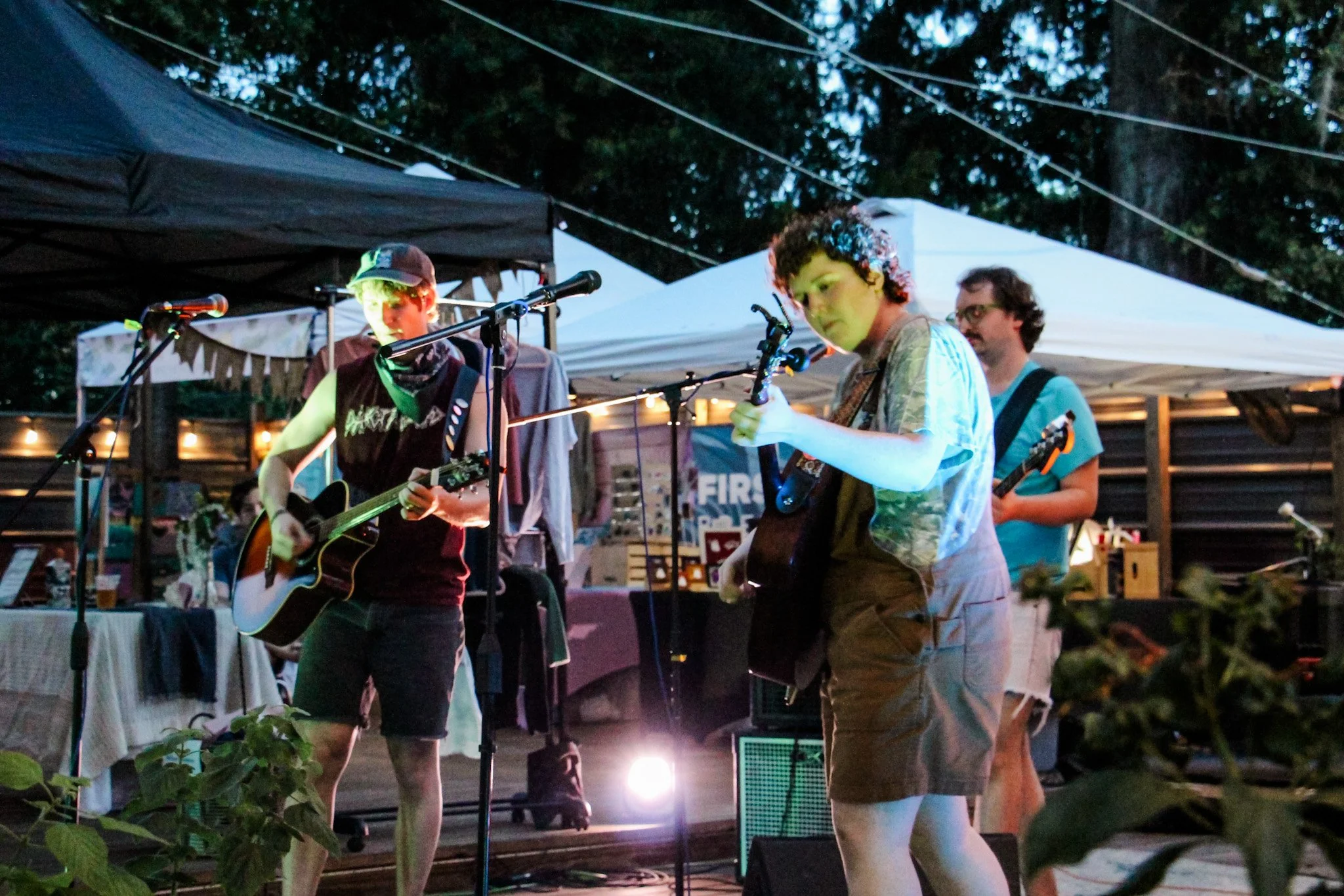 A group of four young men playing guitars at an outdoor event during twilight. There are tents and string lights in the background.